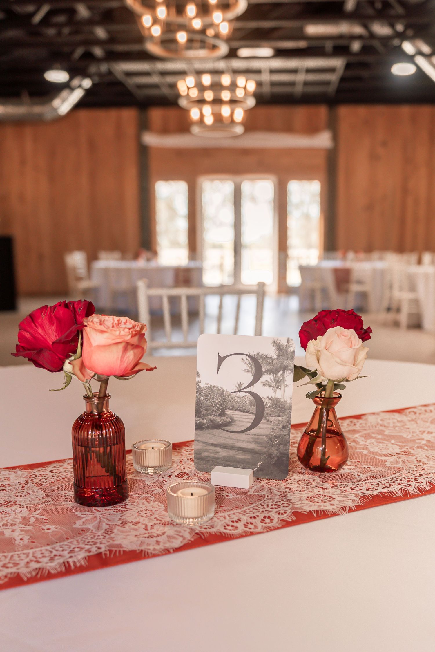 Red and pink floral centerpieces in glass vases line a white tablecloth with lace runner at an elegant indoor venue.