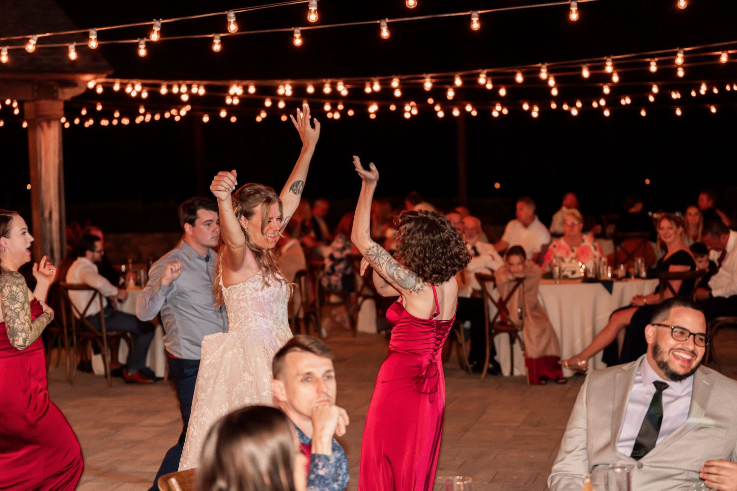 Wedding guests dance and celebrate under glowing string lights at an outdoor evening reception venue.