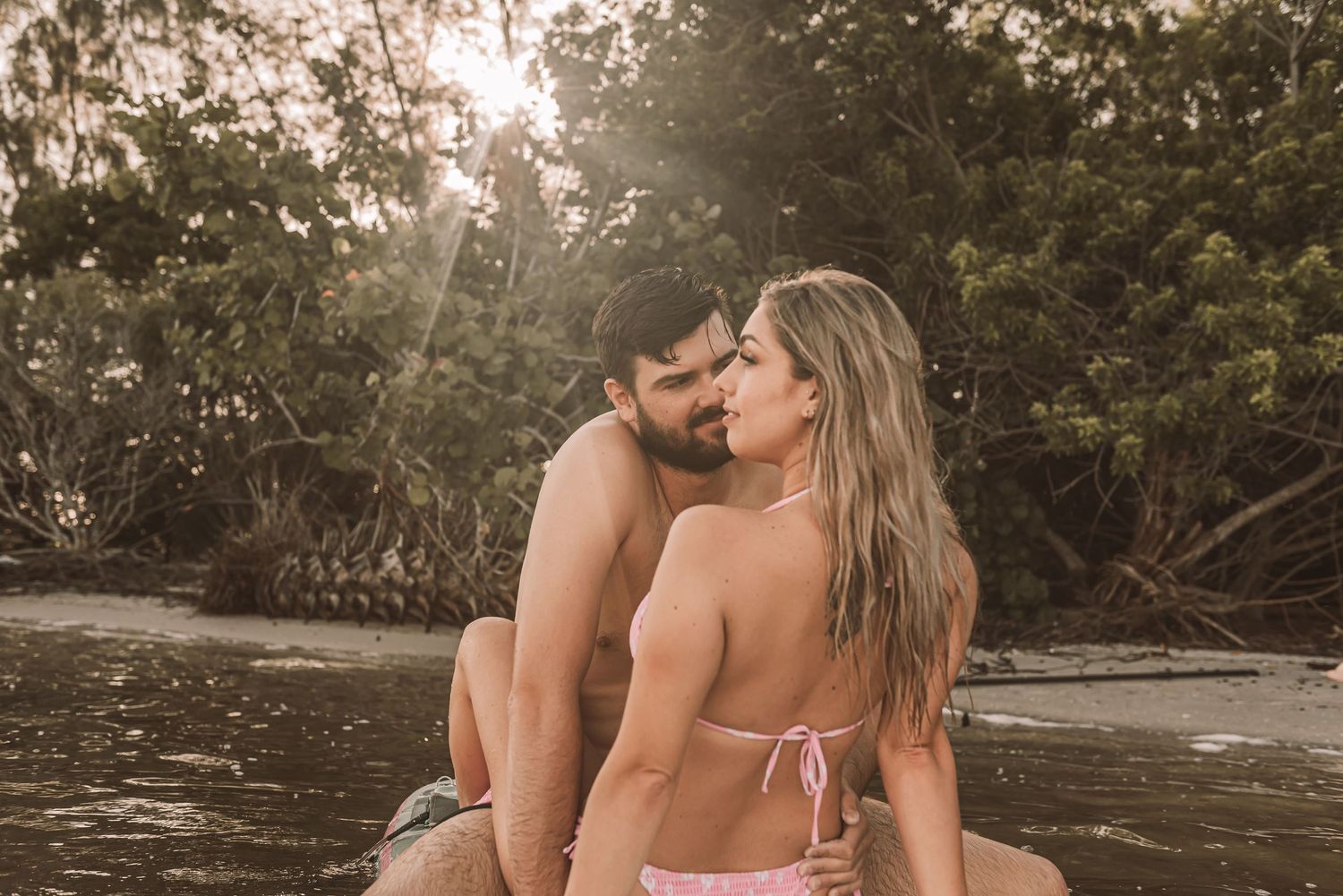 Romantic couple sharing an intimate moment on a rocky beach surrounded by lush tropical trees in golden sunlight.