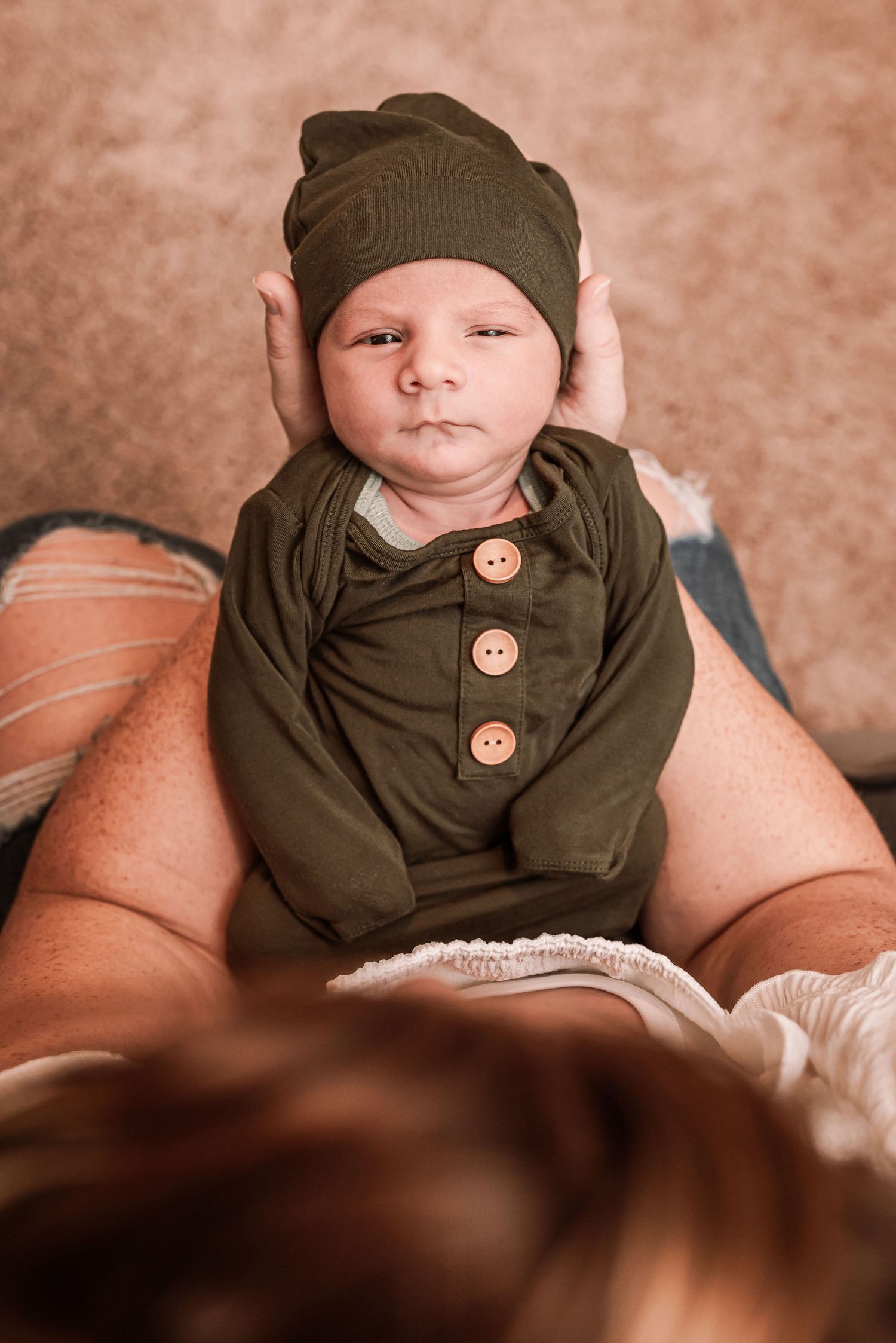 Newborn baby in olive green outfit and matching hat resting on mother's lap during professional photo session.