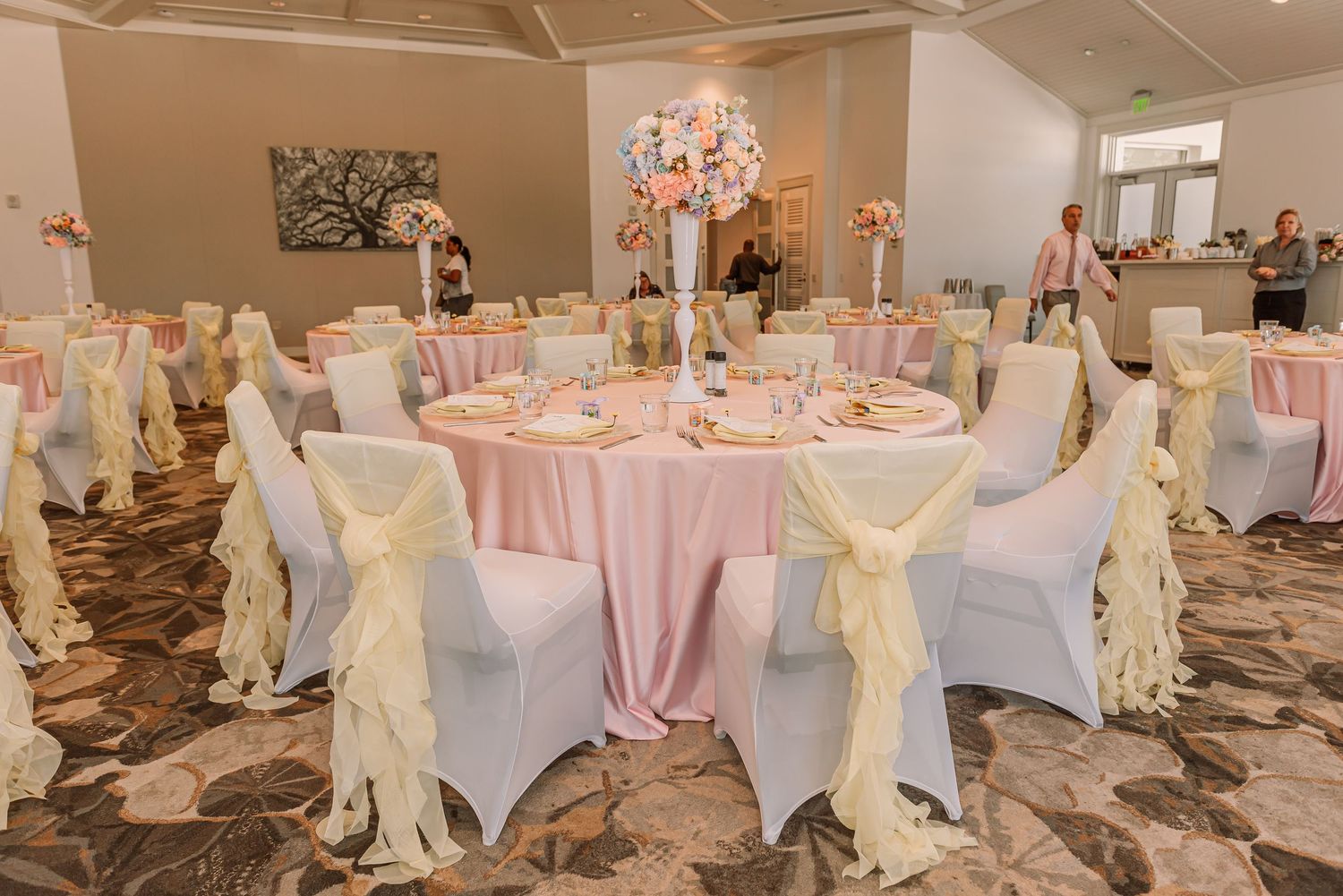 Wedding reception setup with pink tablecloths, white chair covers and gold sashes in an elegant ballroom.