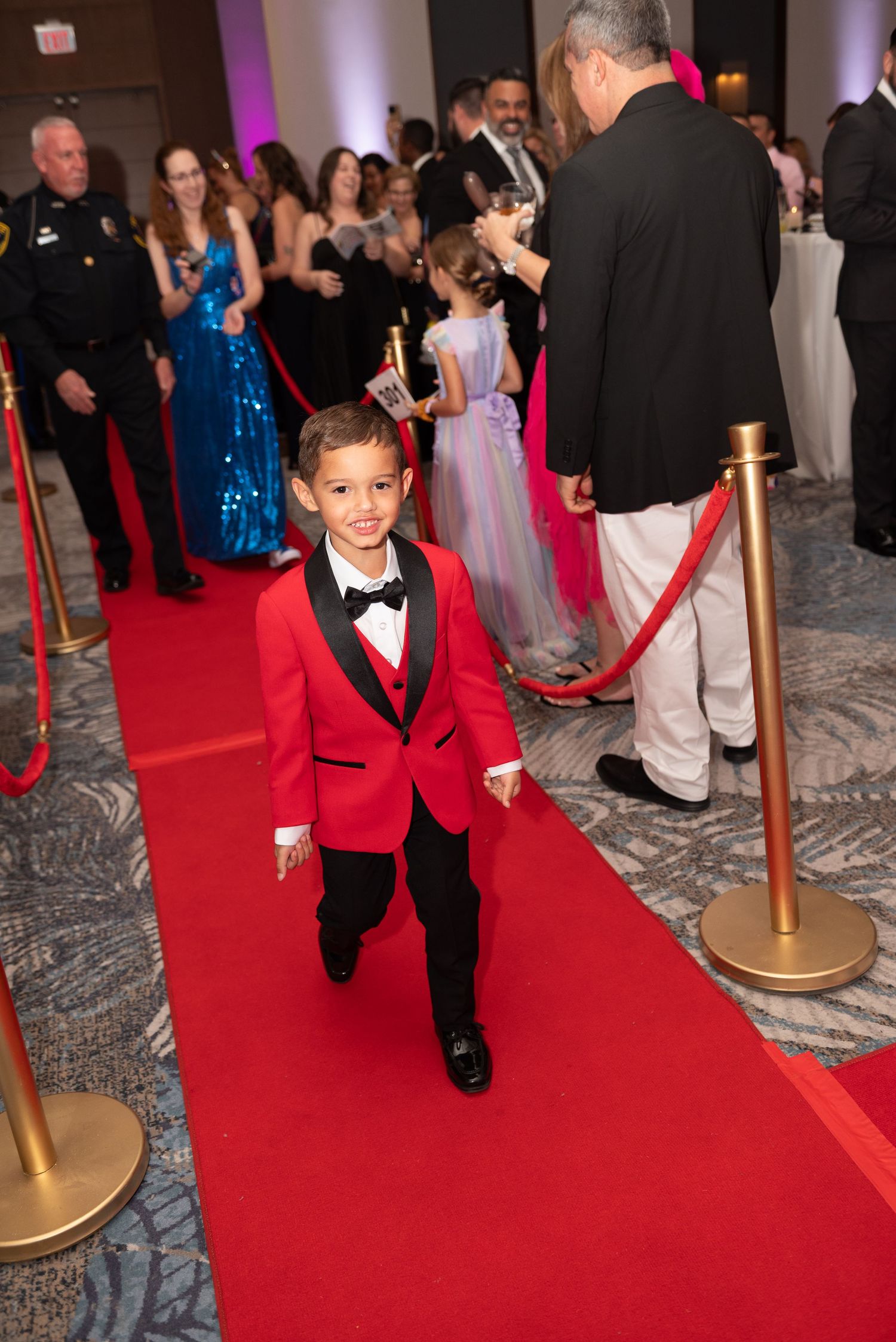 A young boy in a tuxedo runs excitedly down a red carpet at a formal event.