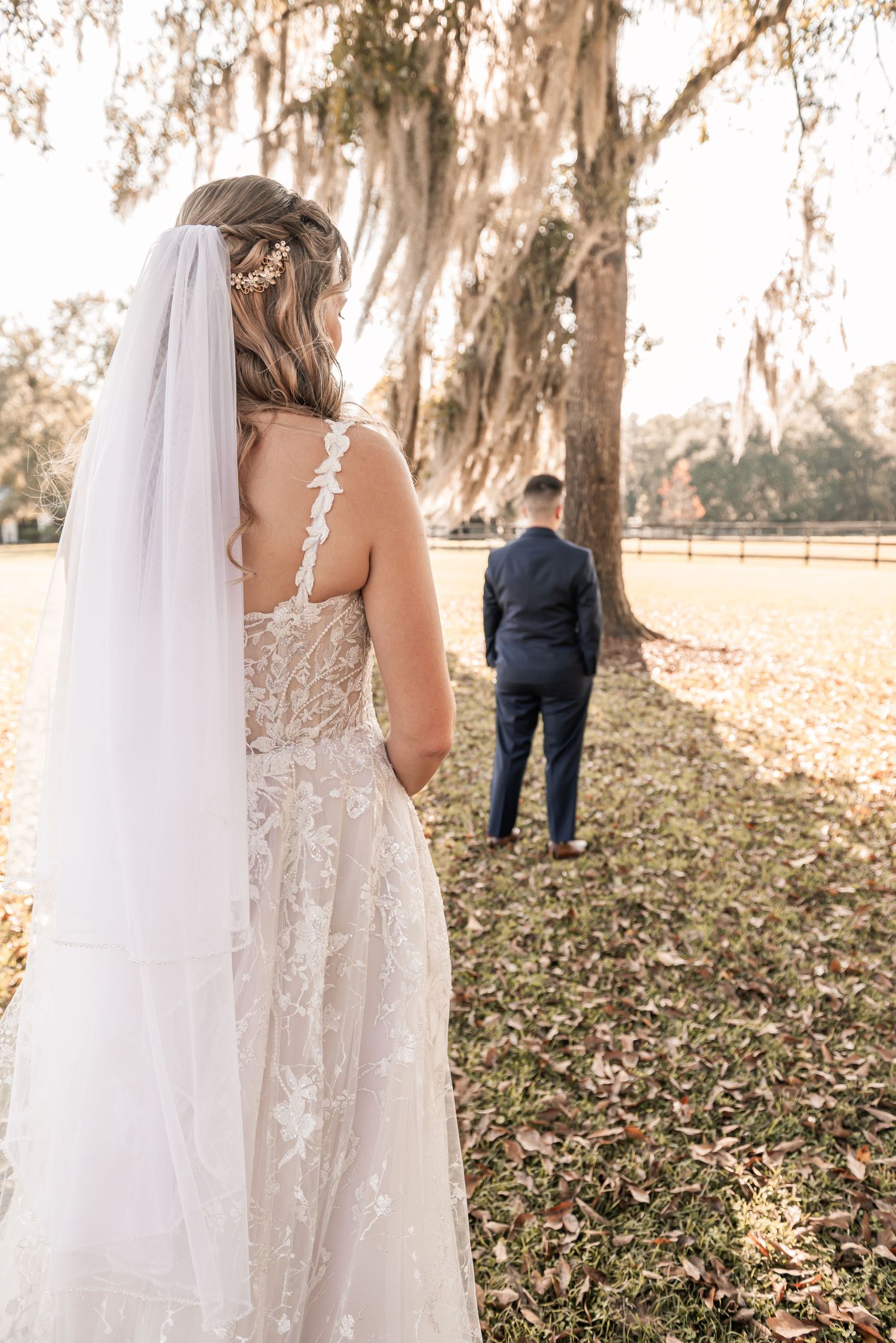 A bride in a lace wedding dress and flowing veil stands among Spanish moss-draped trees during a first look moment.