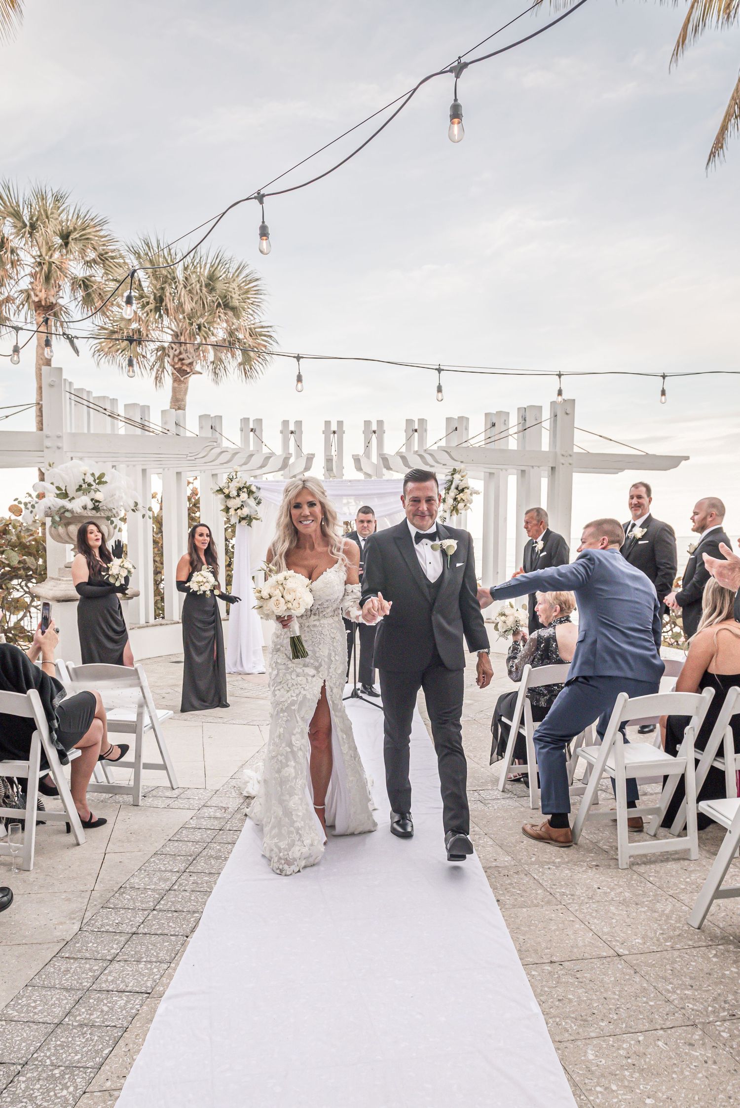 Wedding guests dance and celebrate on an outdoor patio decorated with string lights and palm trees at a beachfront venue.