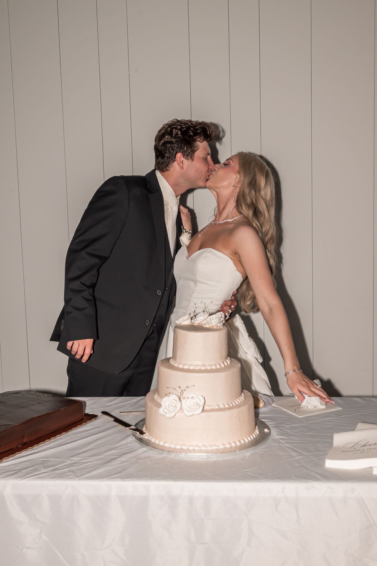 Bride and groom sharing a kiss behind a three-tier white wedding cake at their reception.