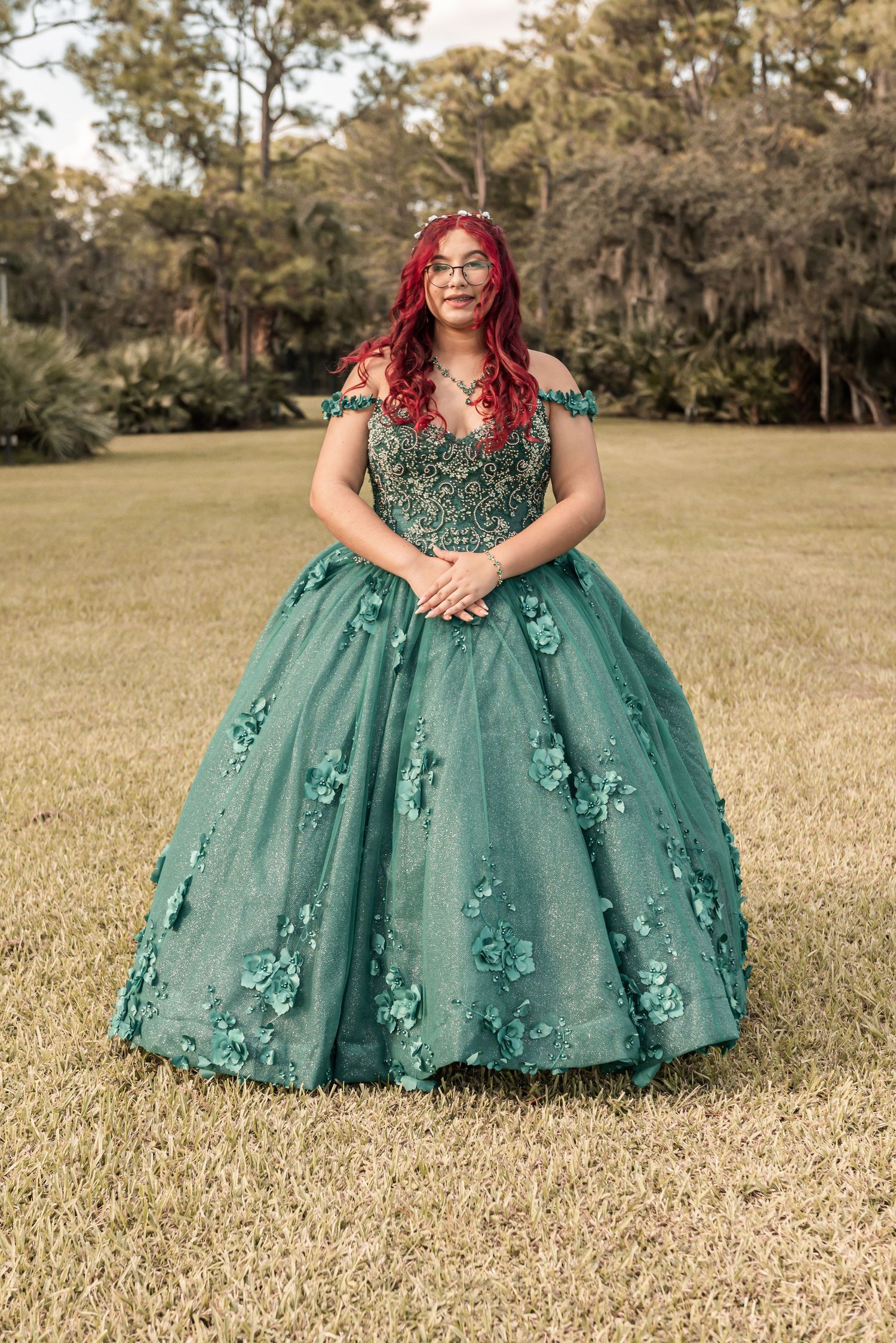 Young woman in elegant green ball gown with floral appliques and red hair poses outdoors for quinceañera photos.