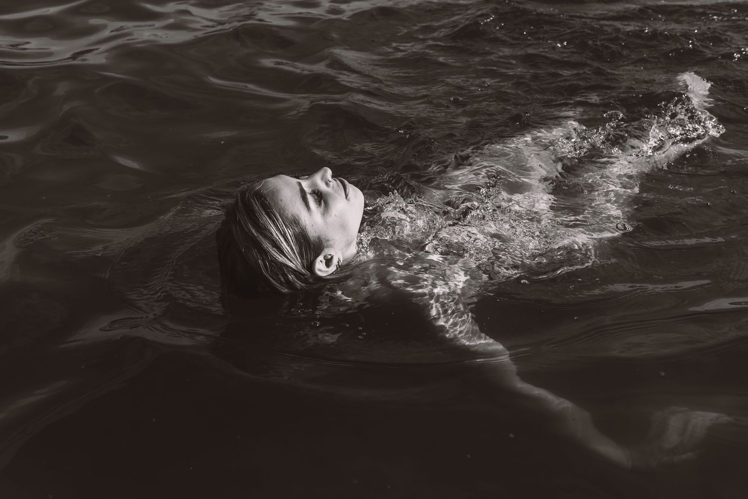 Black and white photo of a person swimming in dark water, head tilted back above the surface.