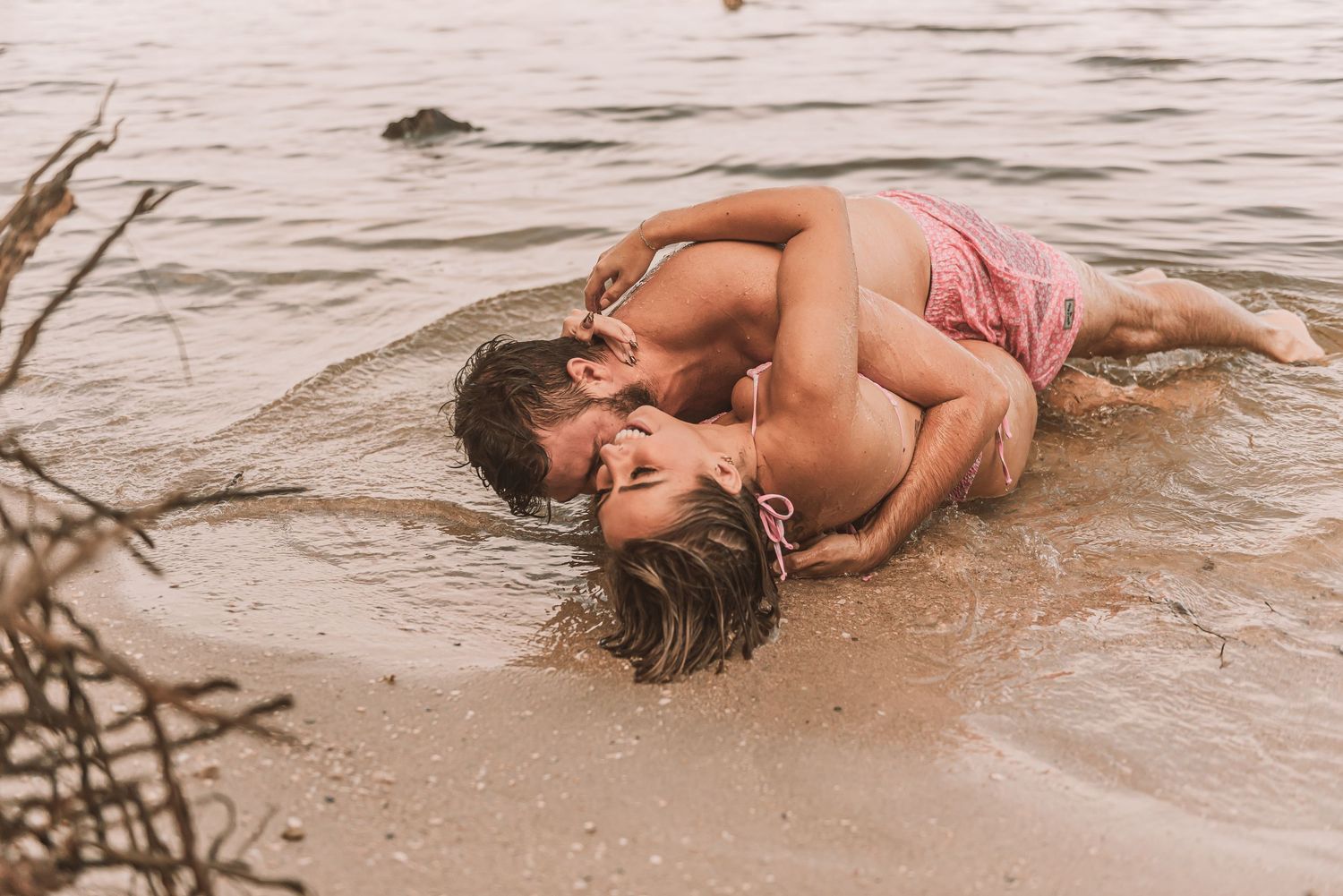 Romantic couple embracing and kissing while lying in shallow ocean waves on a sandy beach.