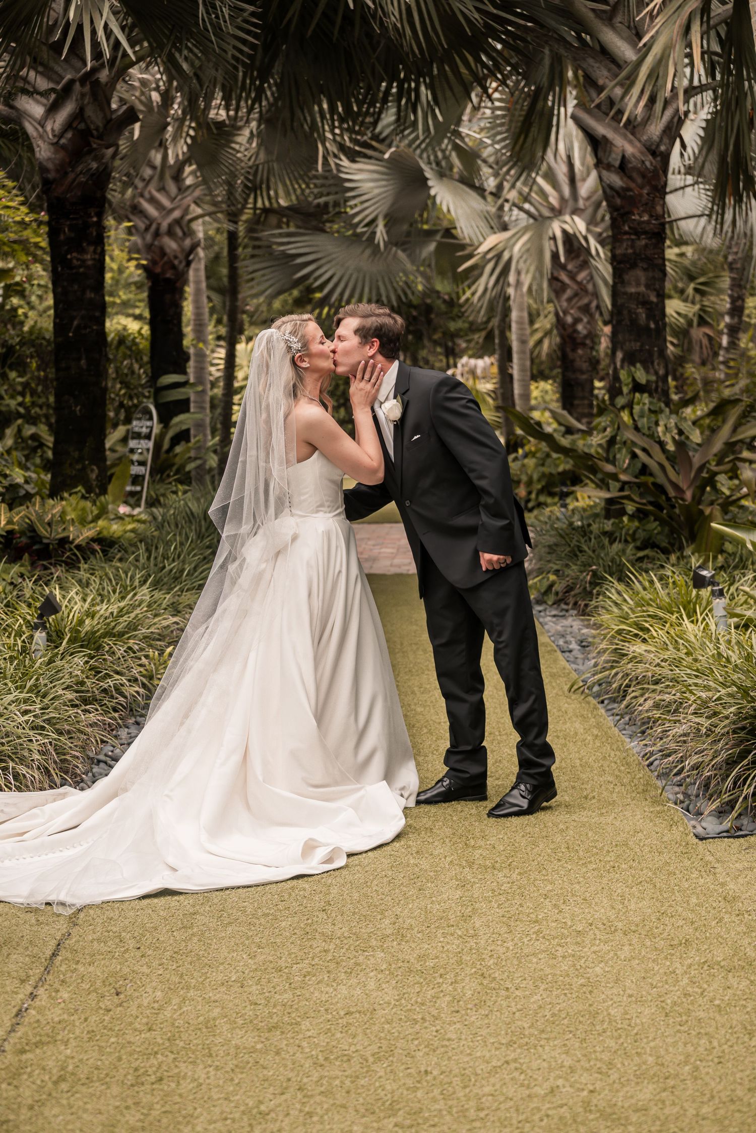 Bride and groom share a romantic kiss on a garden path surrounded by lush palm trees in wedding attire.