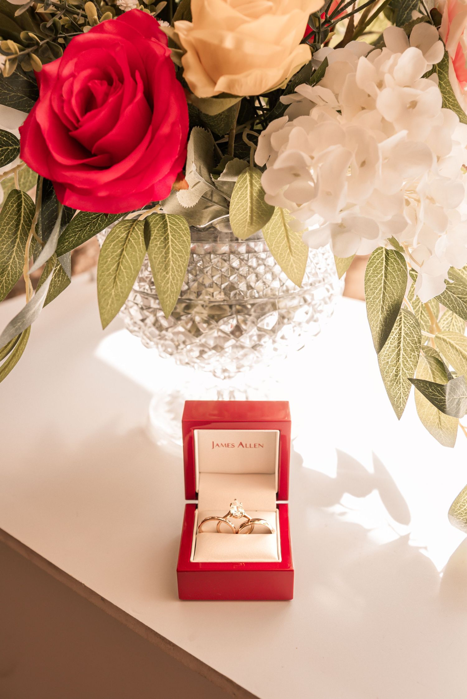 Diamond engagement ring in red James Allen box beside crystal vase with red and white roses.