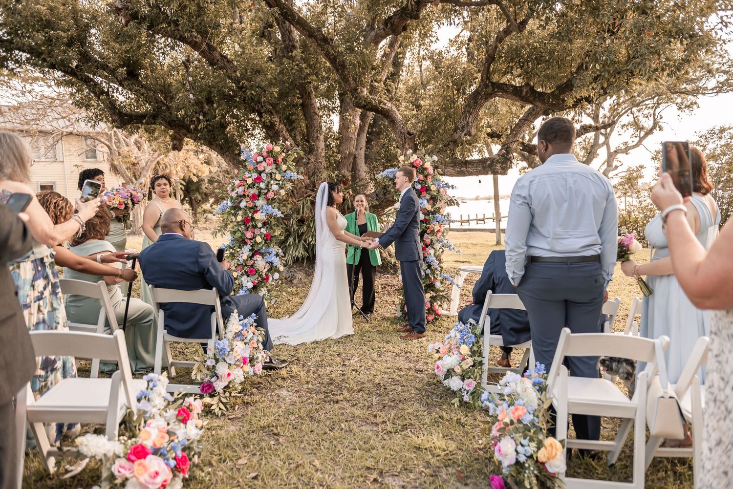 Outdoor wedding ceremony under oak tree with colorful floral aisle, guests seated in white chairs, bride and groom at altar.