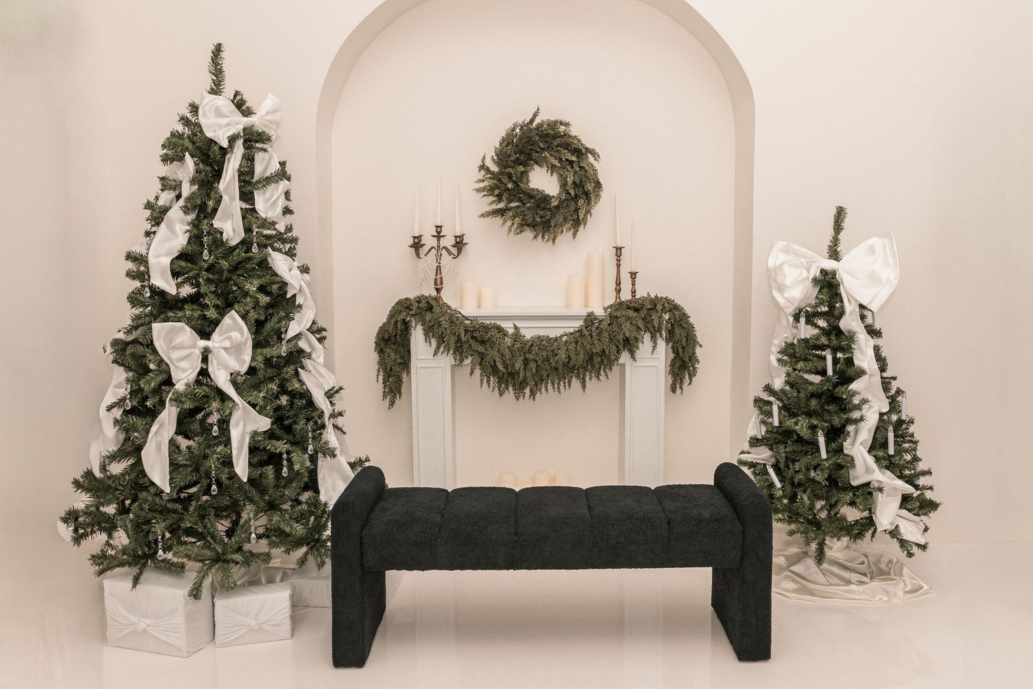 Elegant Christmas photo backdrop with two decorated trees, black bench, and evergreen wreath and garland against white wall.