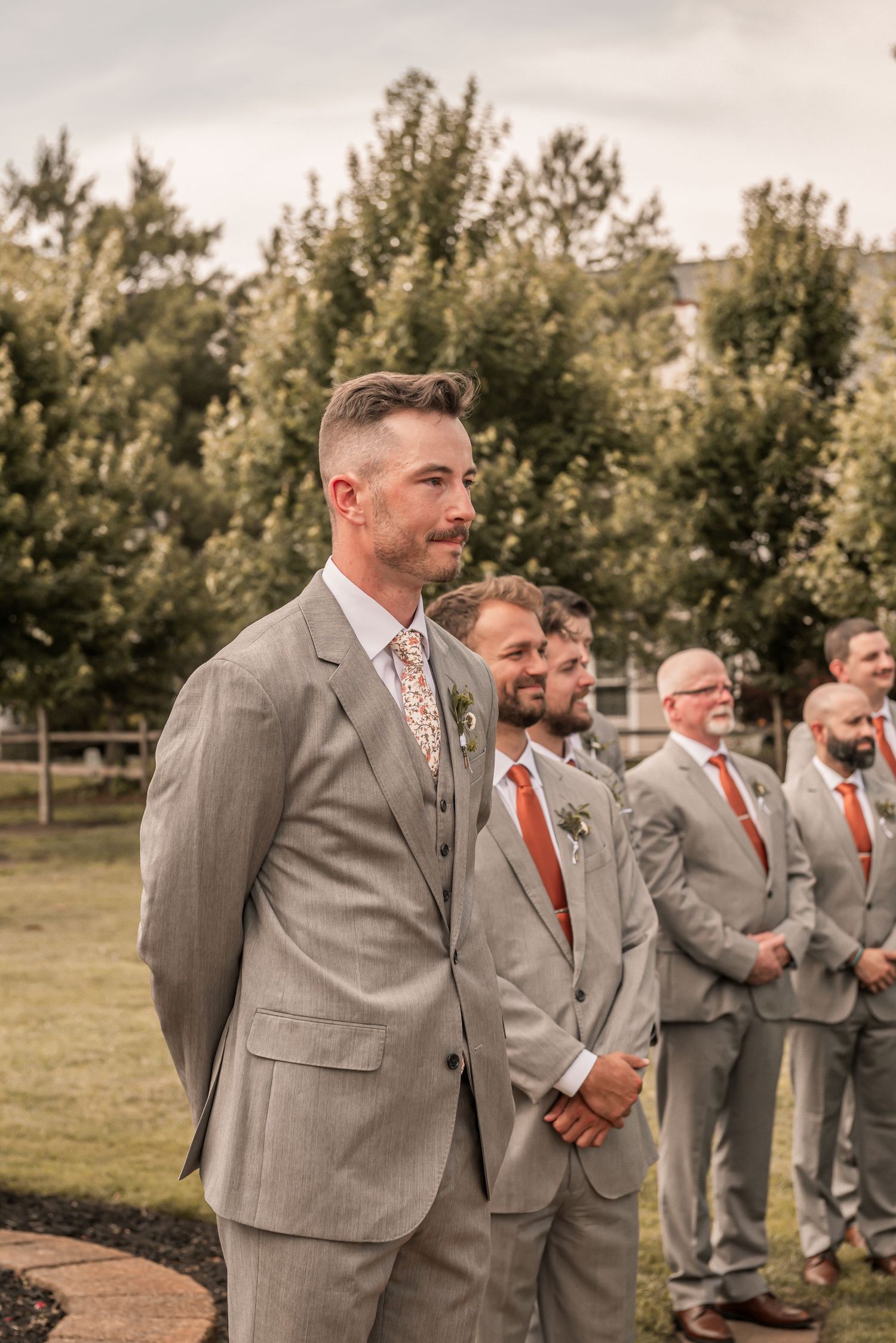 Groomsmen in gray suits and orange ties share a laugh while standing in a line outdoors during a wedding.