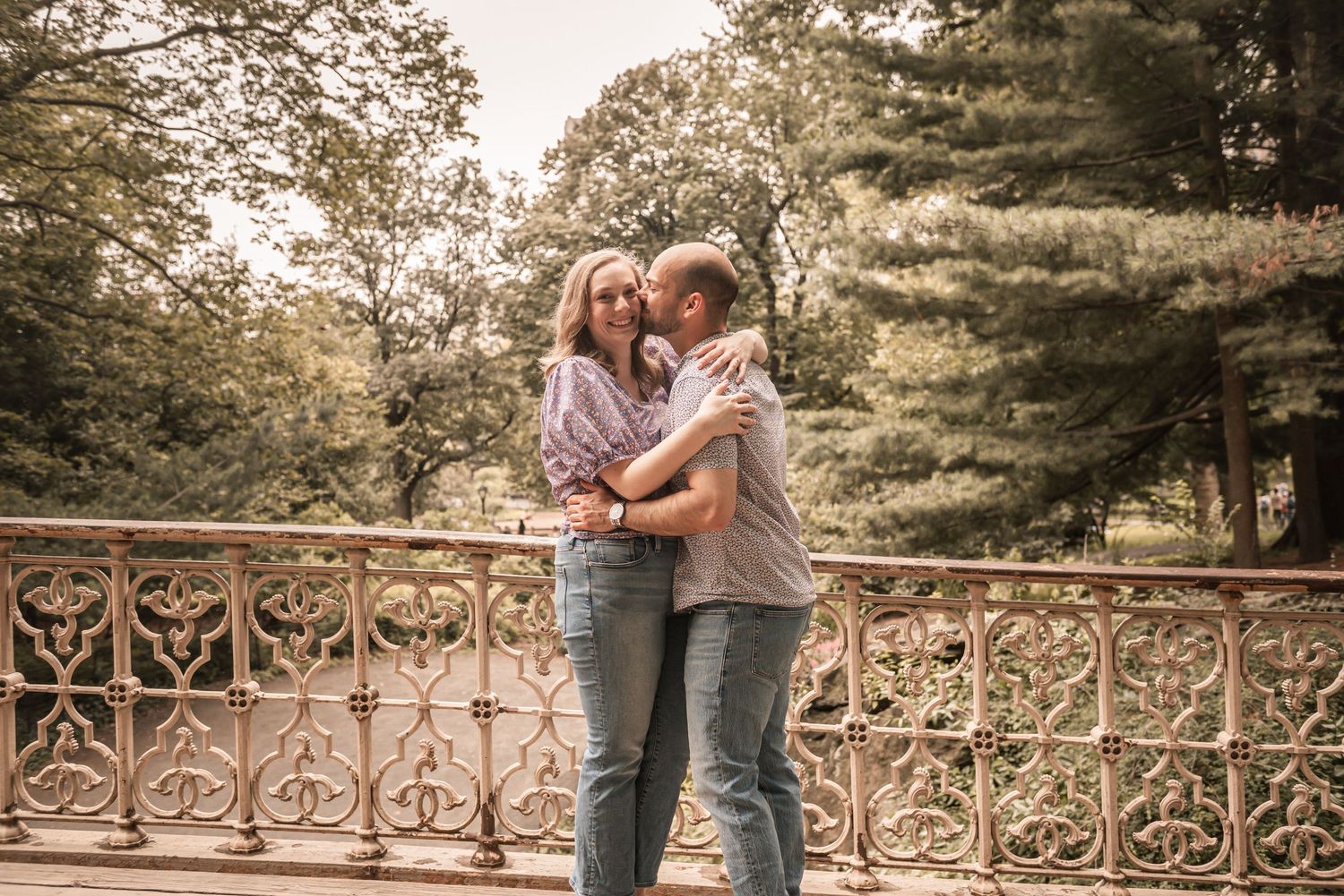 A romantic couple embrace while standing among trees and ornate iron railings during a sunset photoshoot.