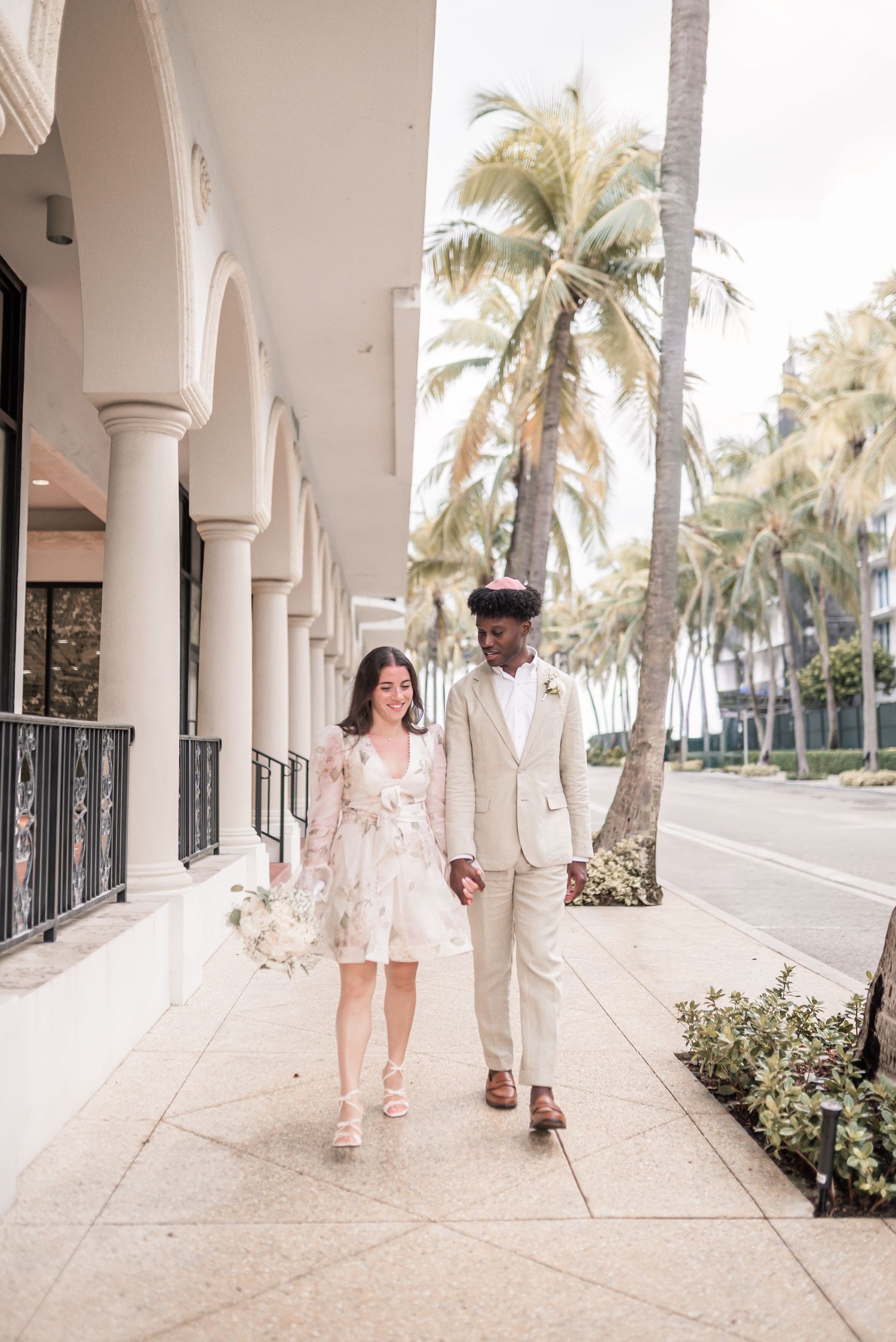 A couple strolls along a palm-lined colonnade with white columns and tropical landscaping.