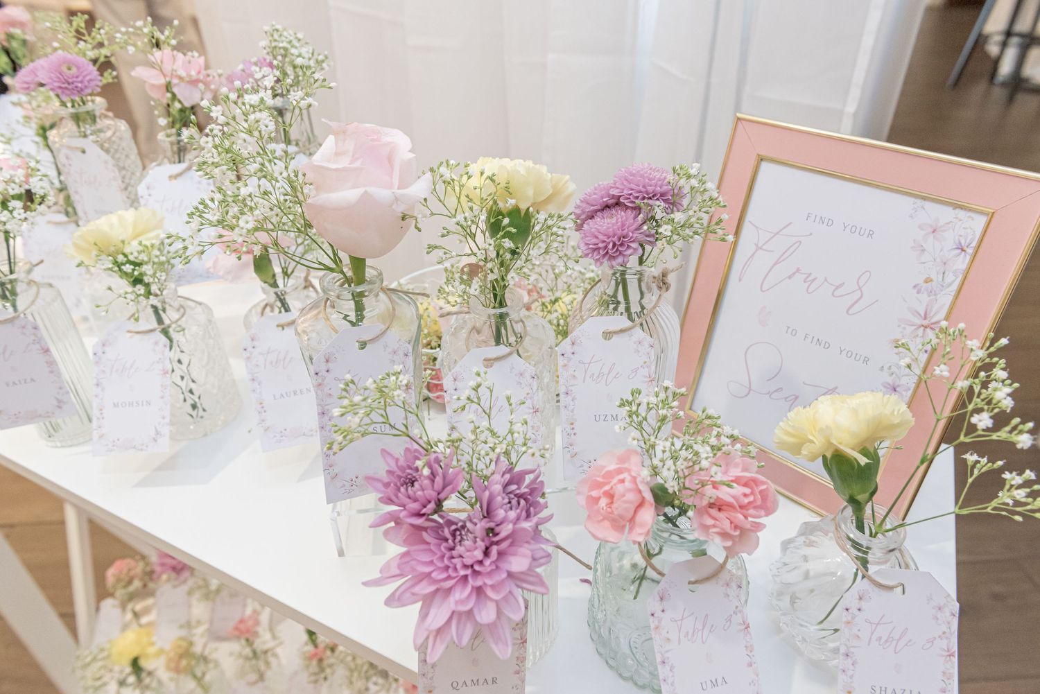 Delicate floral arrangements with pink and purple blooms in glass vases displayed next to a framed sign on a white table.