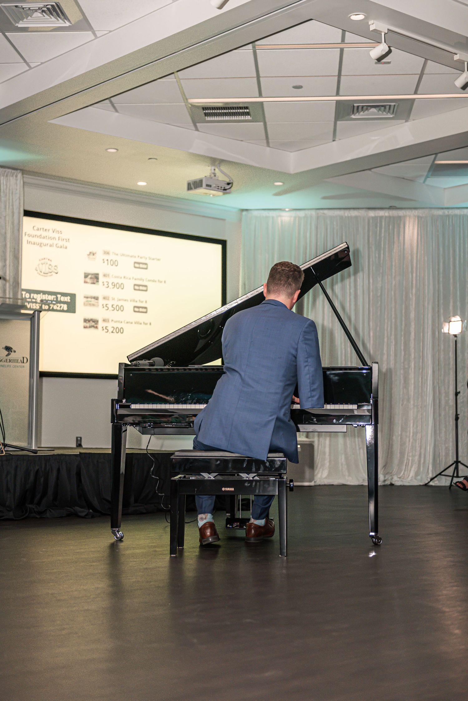 A musician in a blue suit performs at a grand piano on stage with a projector screen and curtains in the background.