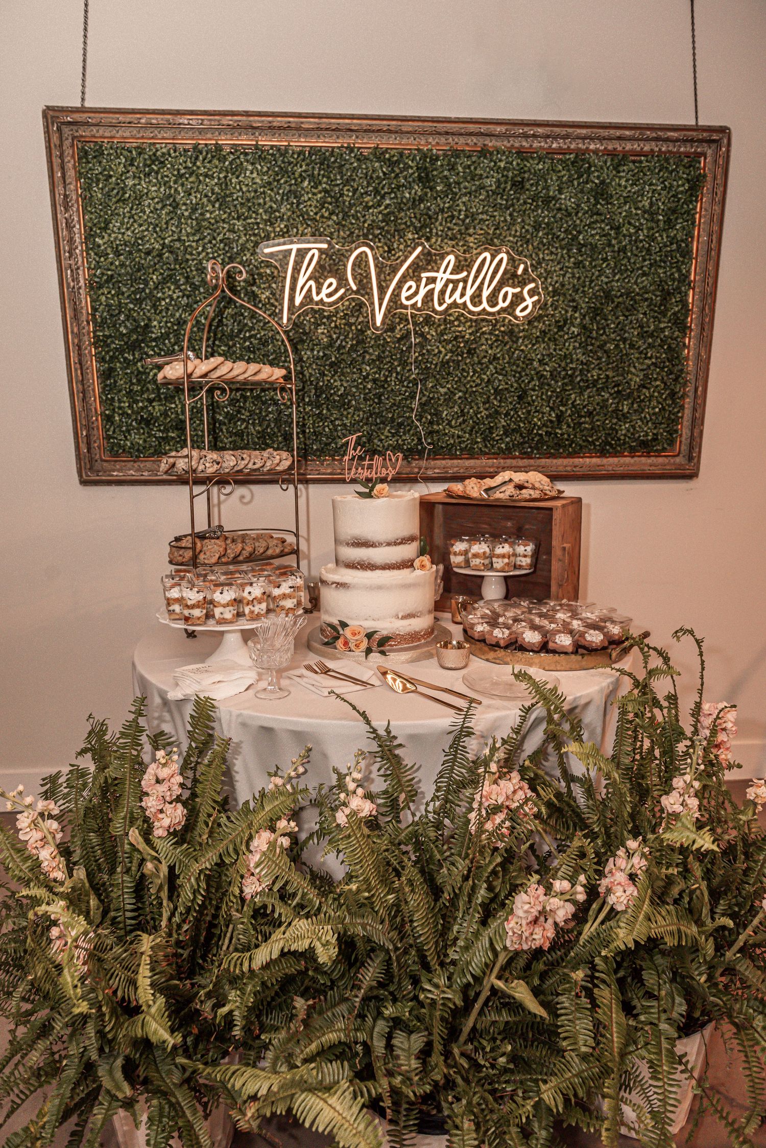 A rustic wedding dessert table with neon sign, white cake, cupcakes and greenery arrangements against a boxwood backdrop.