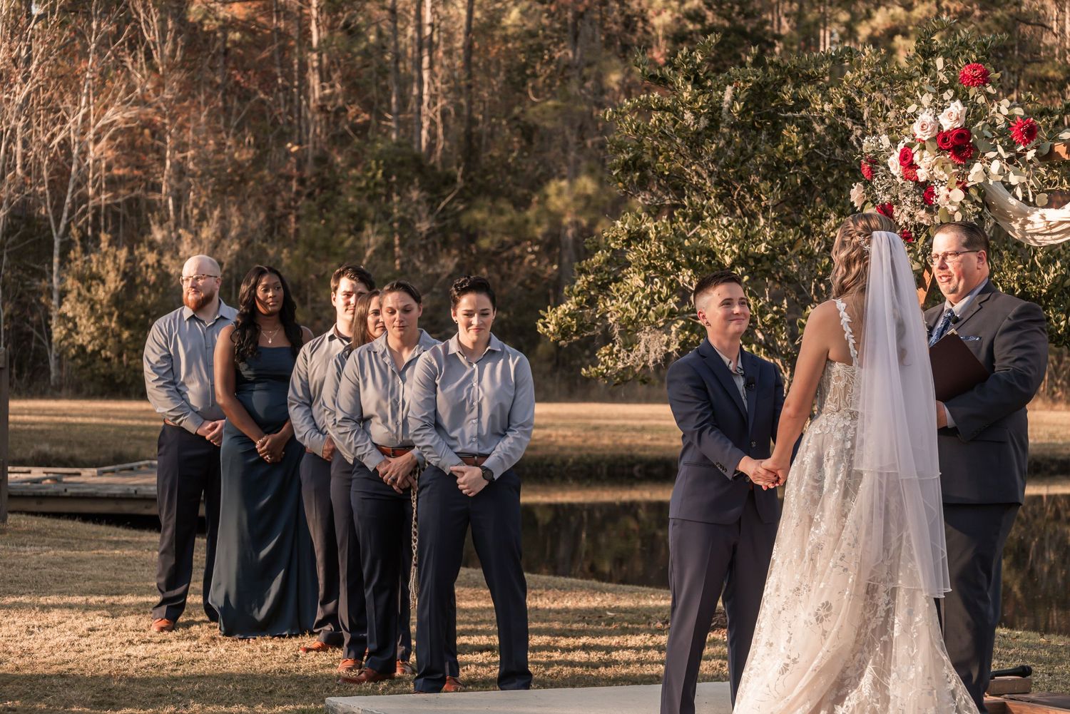 Wedding party in navy suits and white dress poses together outdoors during autumn sunset ceremony.