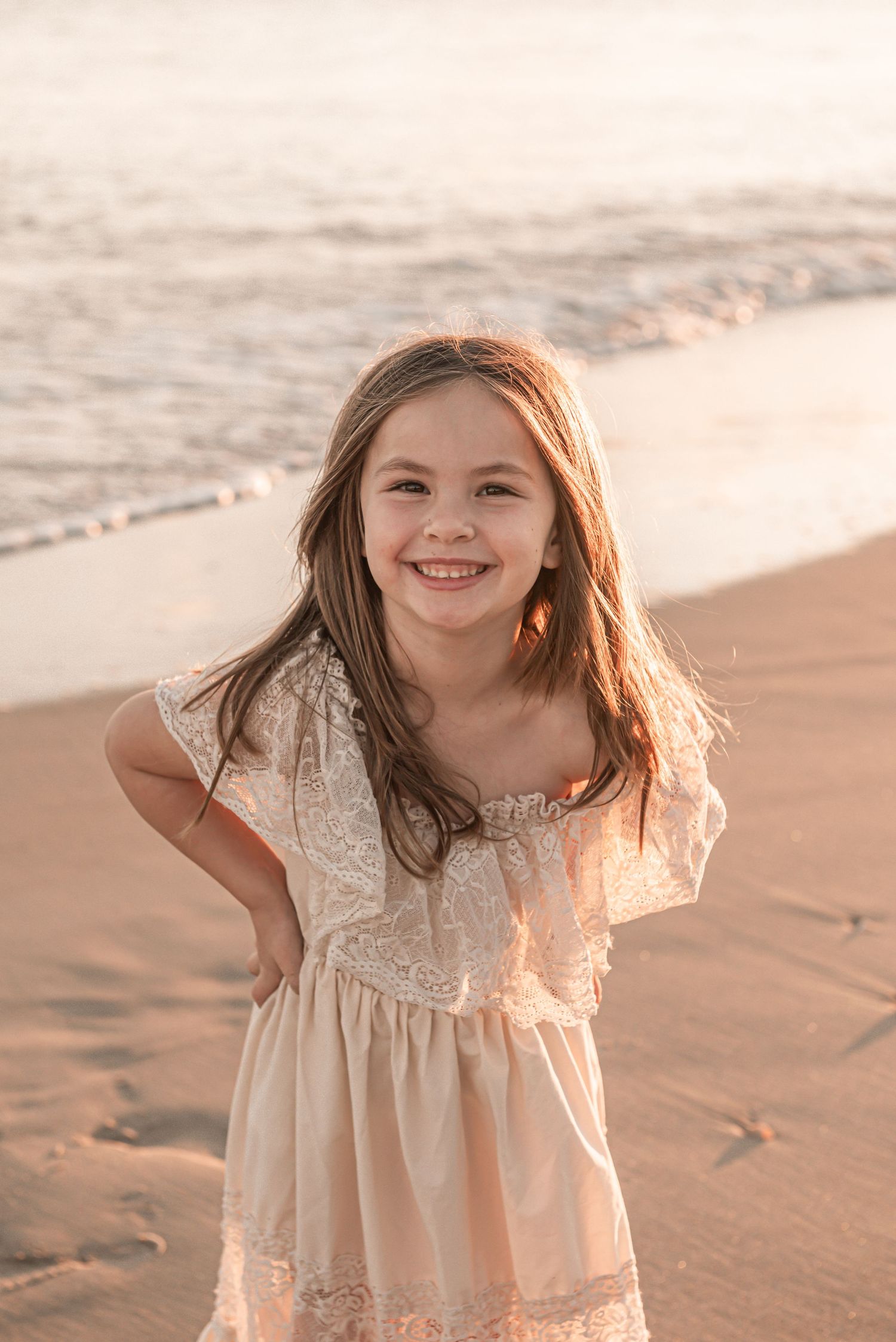 A child in a white lace dress plays joyfully at the beach during golden hour.