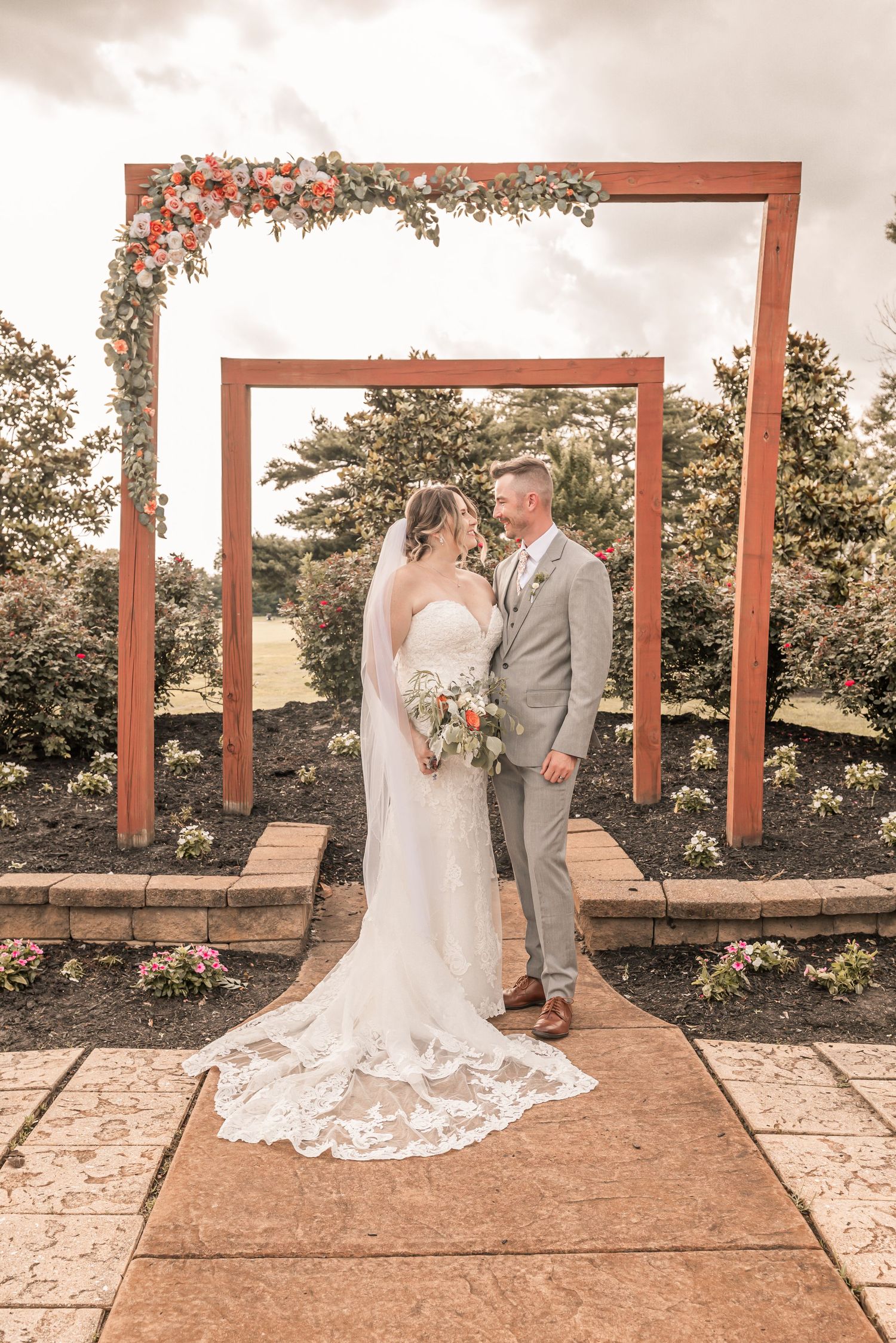 Two figures in wedding attire pose beneath geometric wooden frames decorated with floral arrangements in an outdoor garden setting.