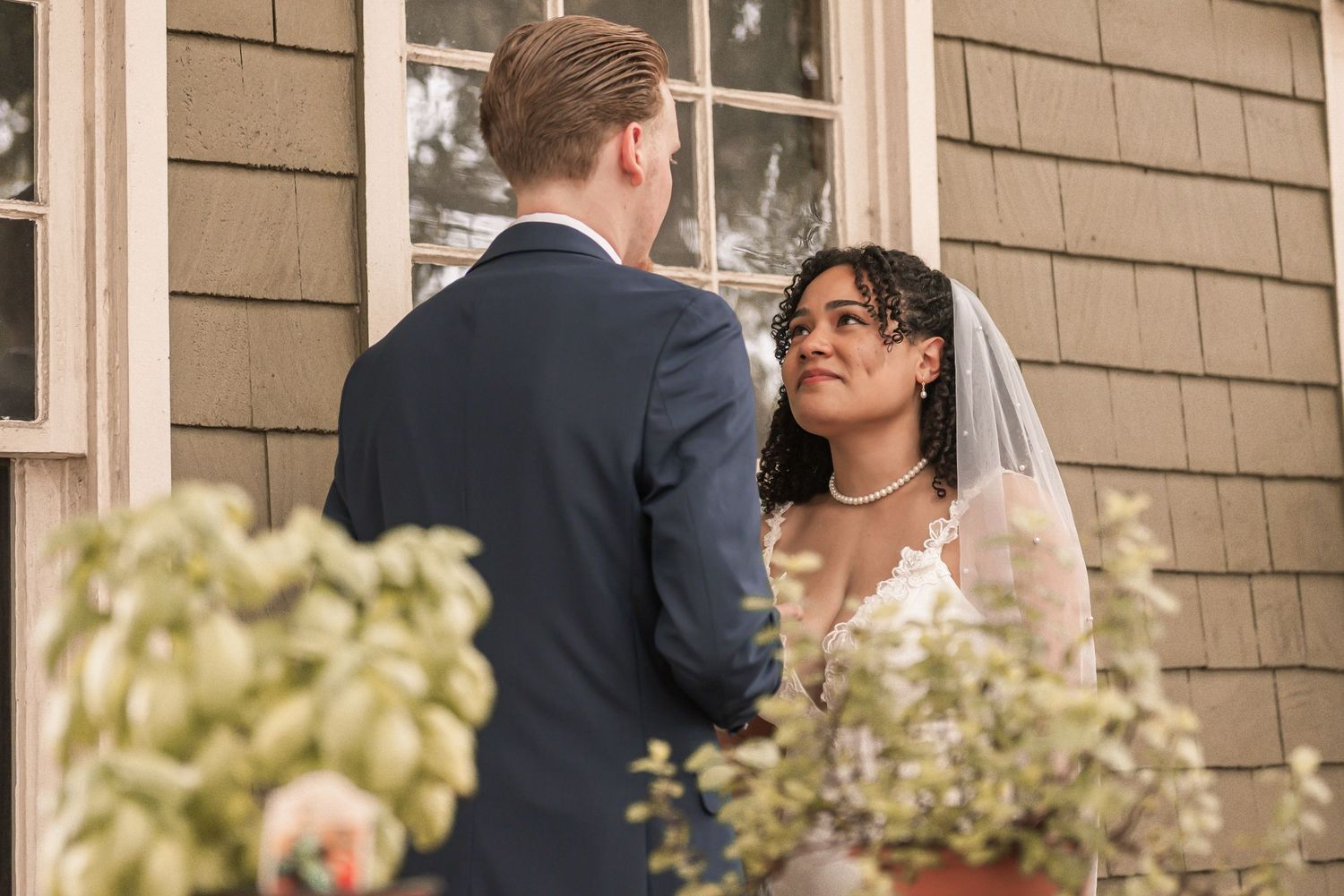Bride in lace gown and veil gazes at groom in navy suit during outdoor wedding ceremony by a wooden building.