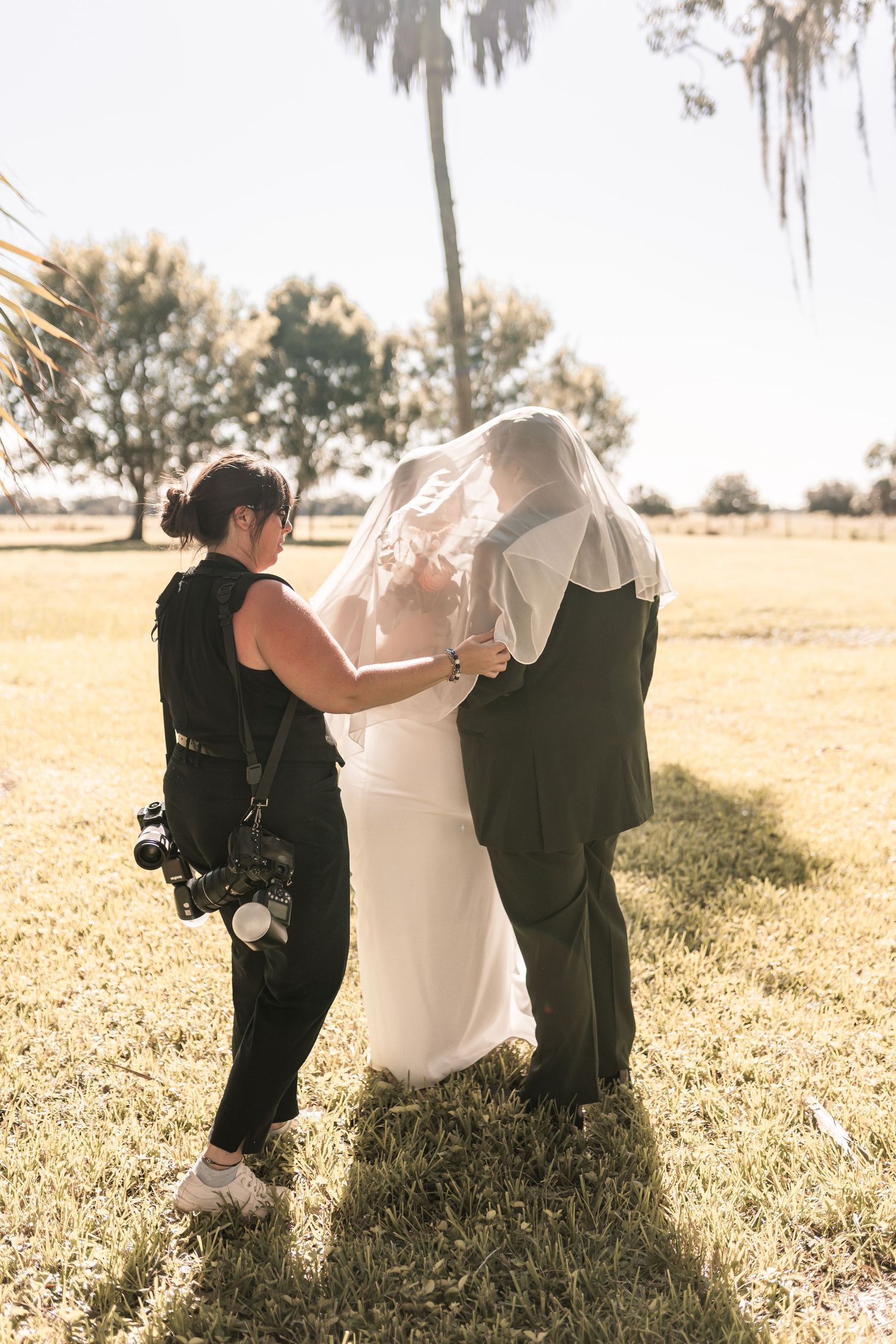 Photographer adjusting bride's veil while couple embraces outdoors on sunny day at wedding venue.
