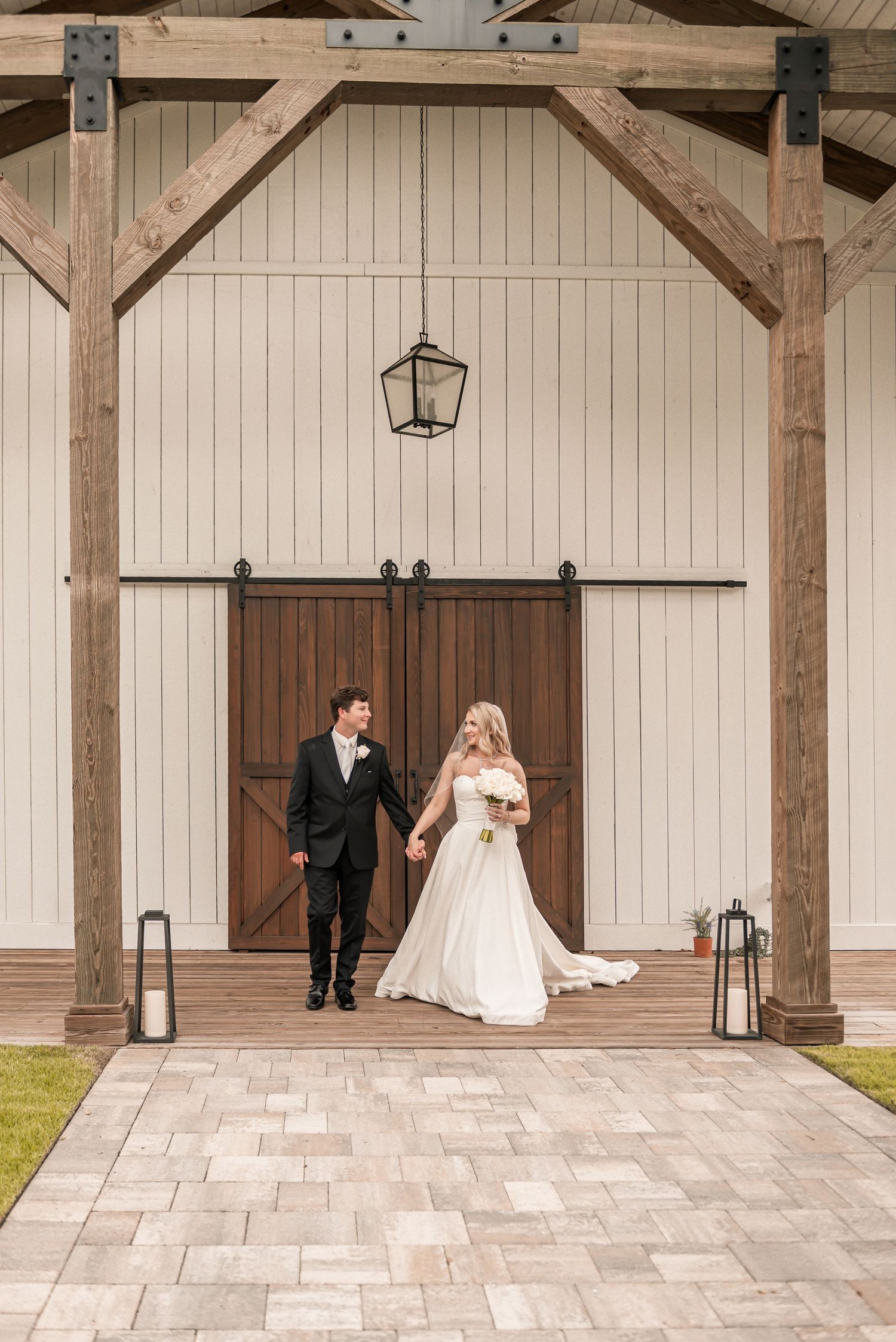 Bride and groom holding hands in front of rustic barn doors with wooden beams and lanterns on a brick pathway.