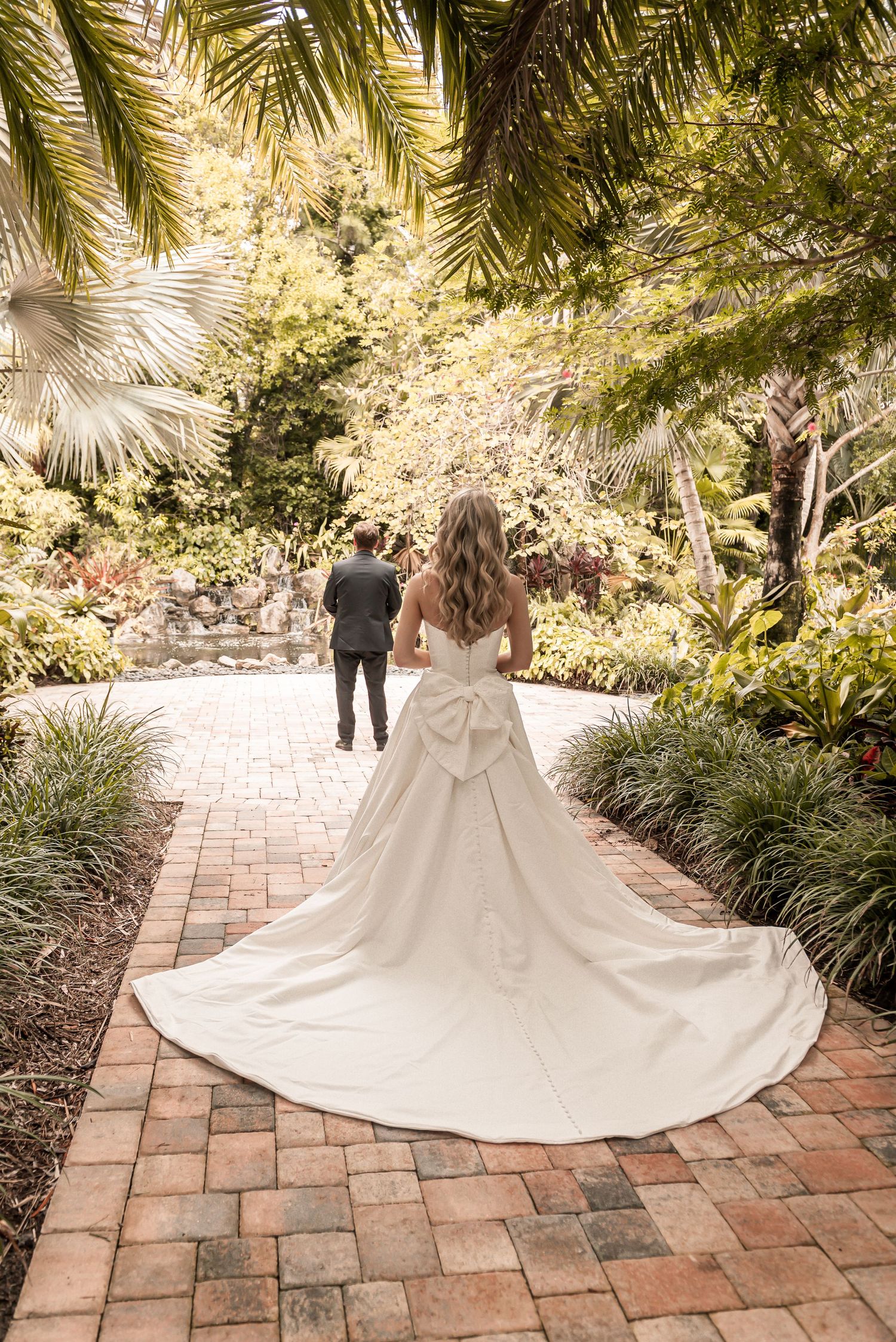 Bride in white gown with large bow train walks toward groom on brick path in lush tropical garden.