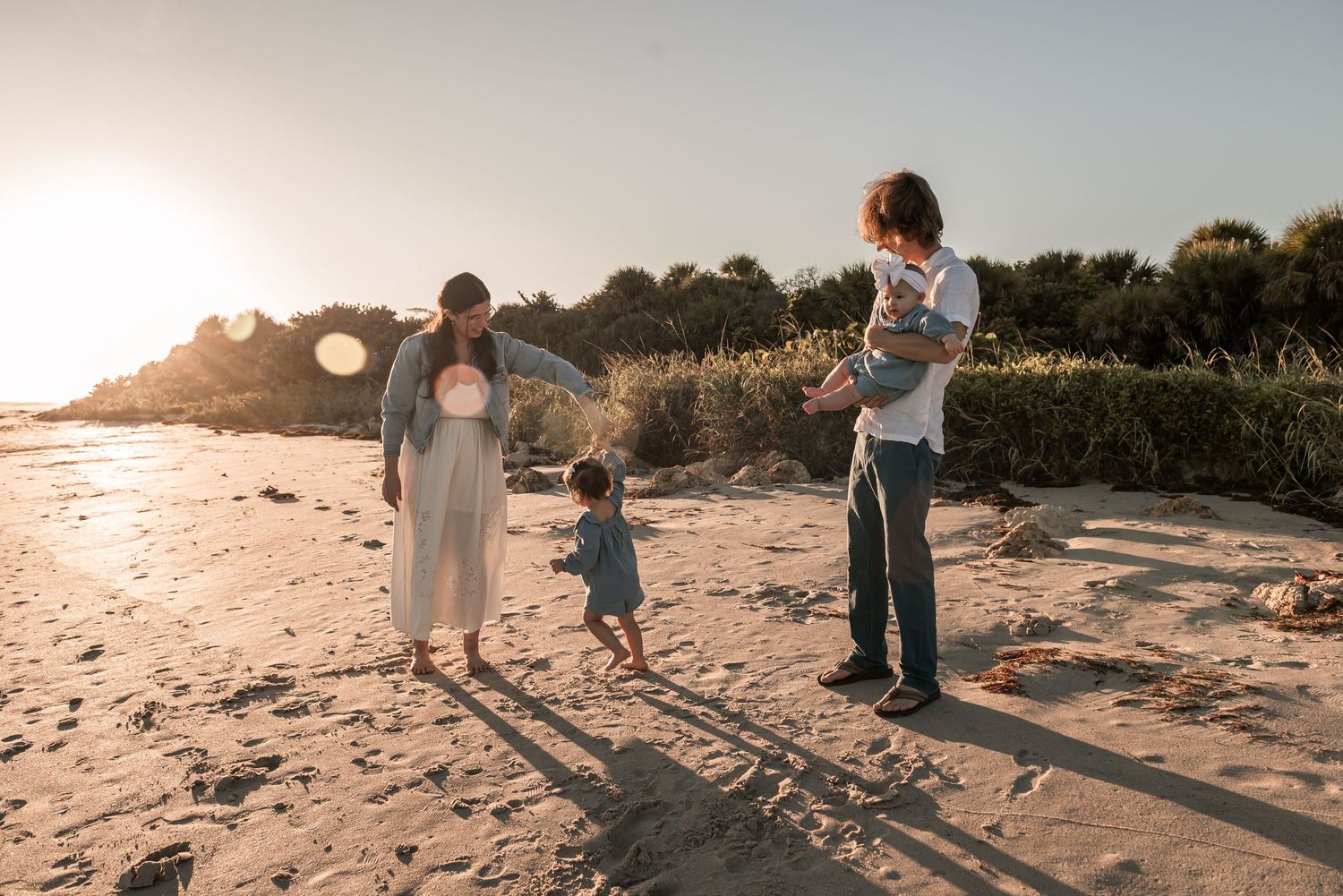 Young family walking on sandy beach at golden sunset with toddler and baby.