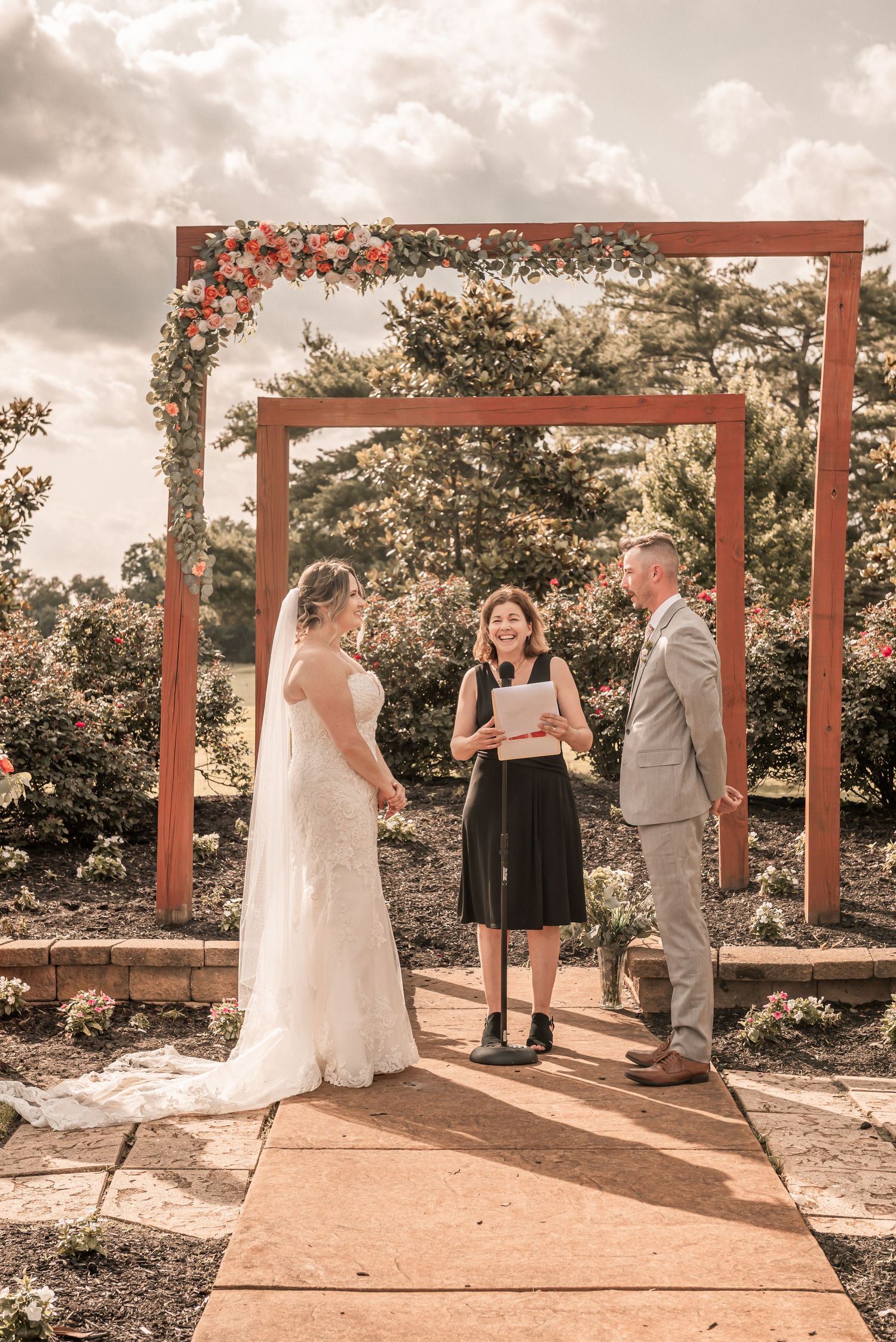 An outdoor wedding ceremony takes place under wooden archways decorated with floral arrangements at sunset.