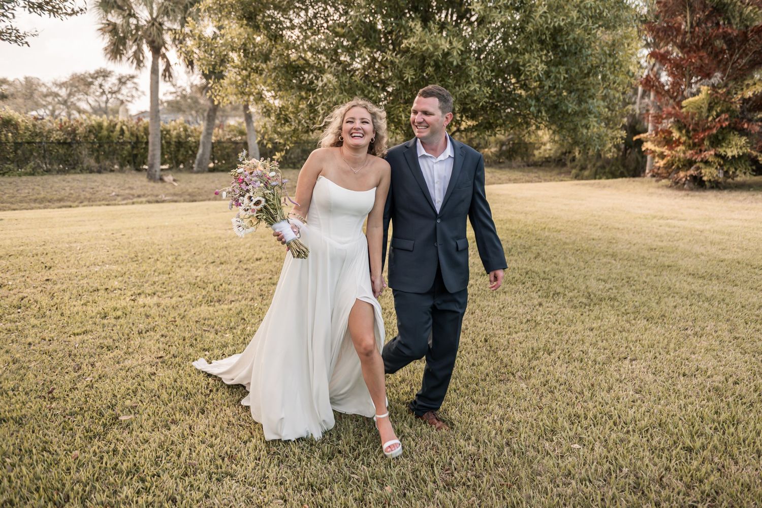 Smiling bride in white gown holding bouquet walks with groom in navy suit across green lawn at outdoor wedding.