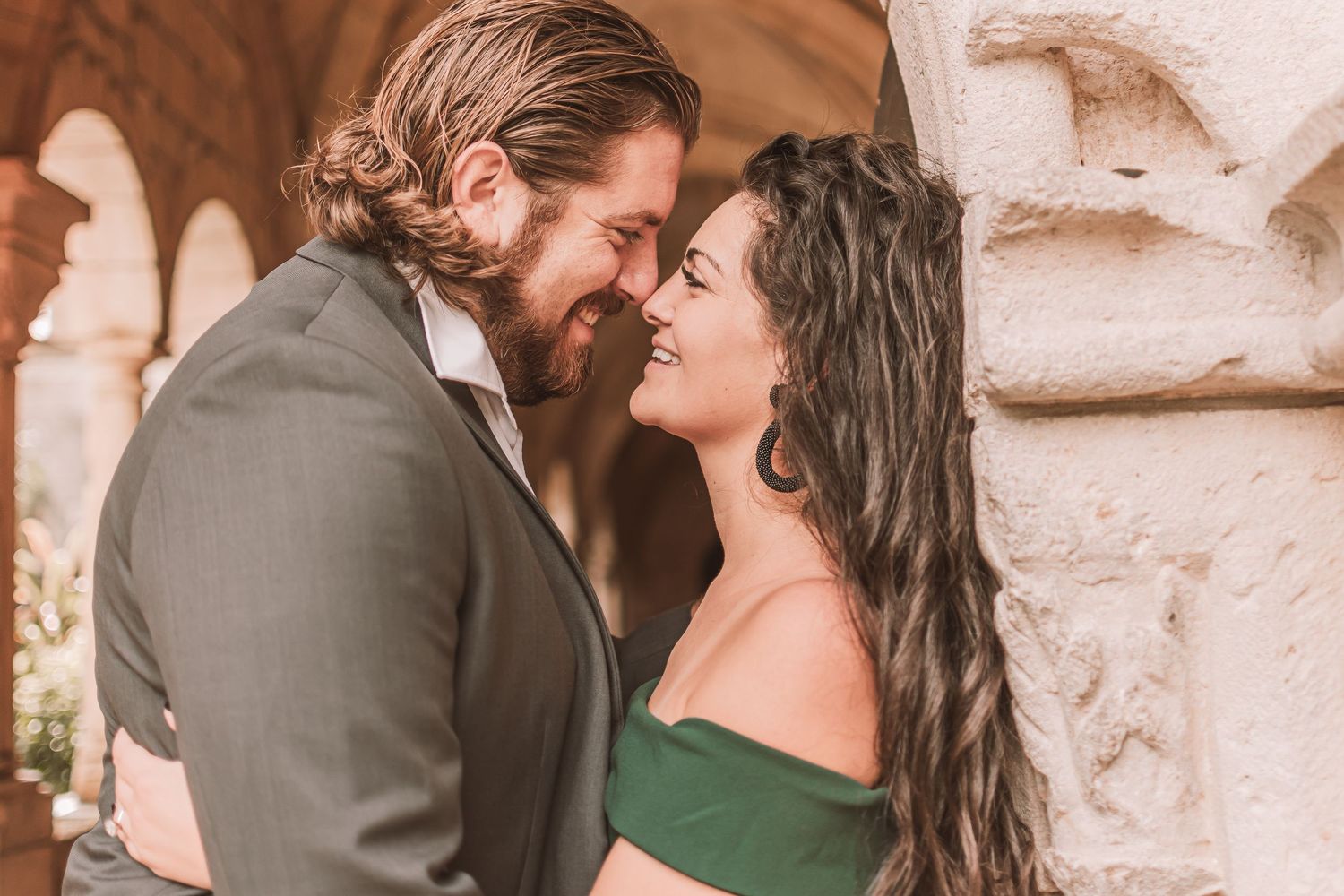 A romantic couple shares an intimate moment while standing close together against a stone wall in warm lighting.