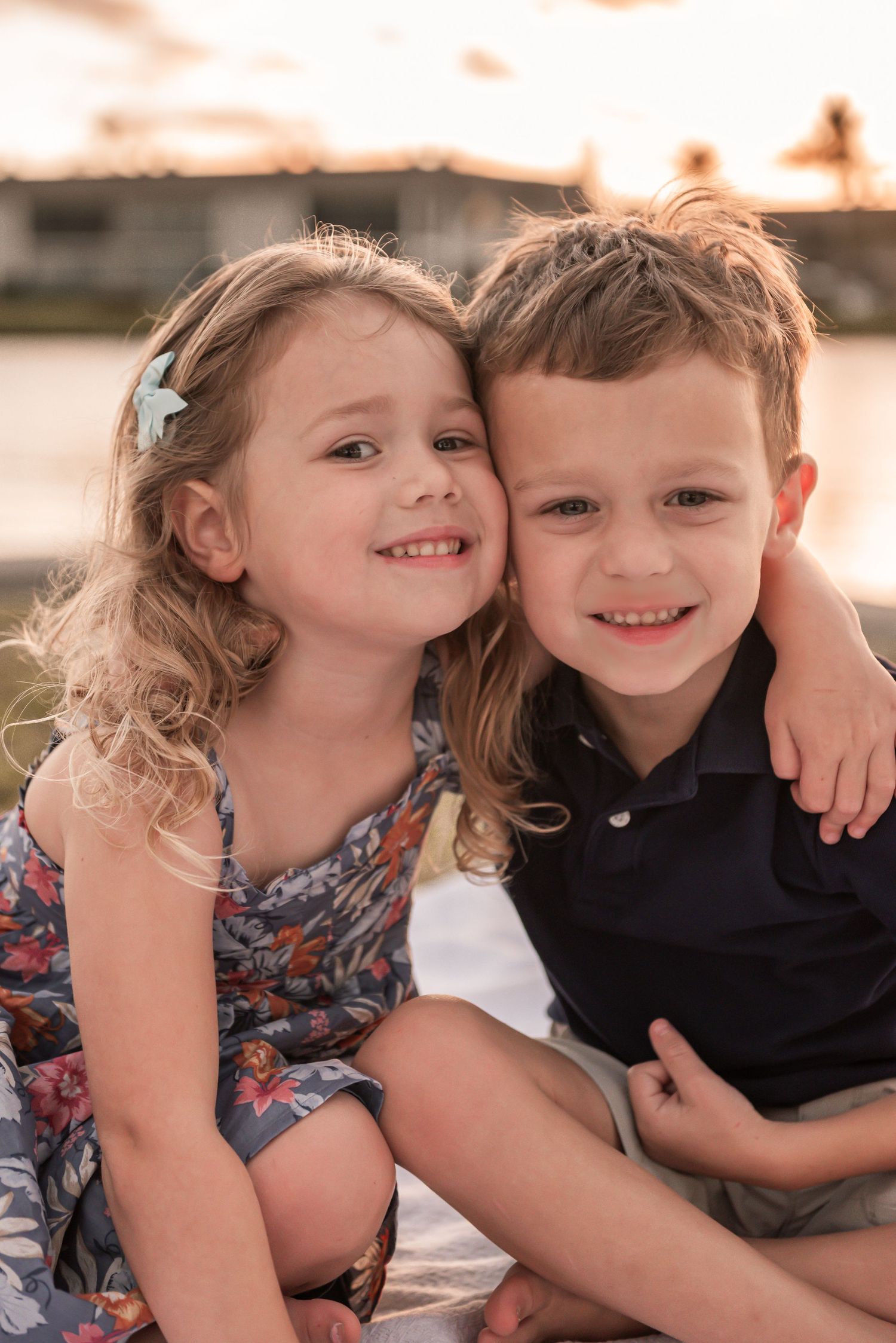 Two young children sit close together outdoors during golden hour wearing casual clothing.