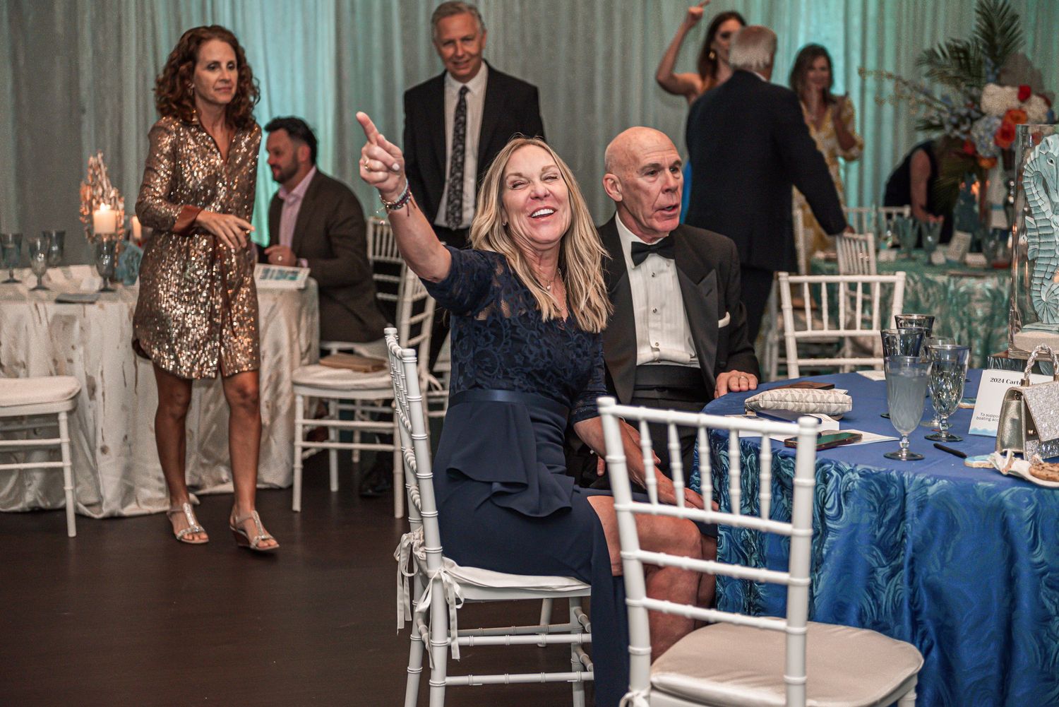 Wedding reception guests in formal attire celebrate at tables with white chairs and blue table linens in an elegant venue.