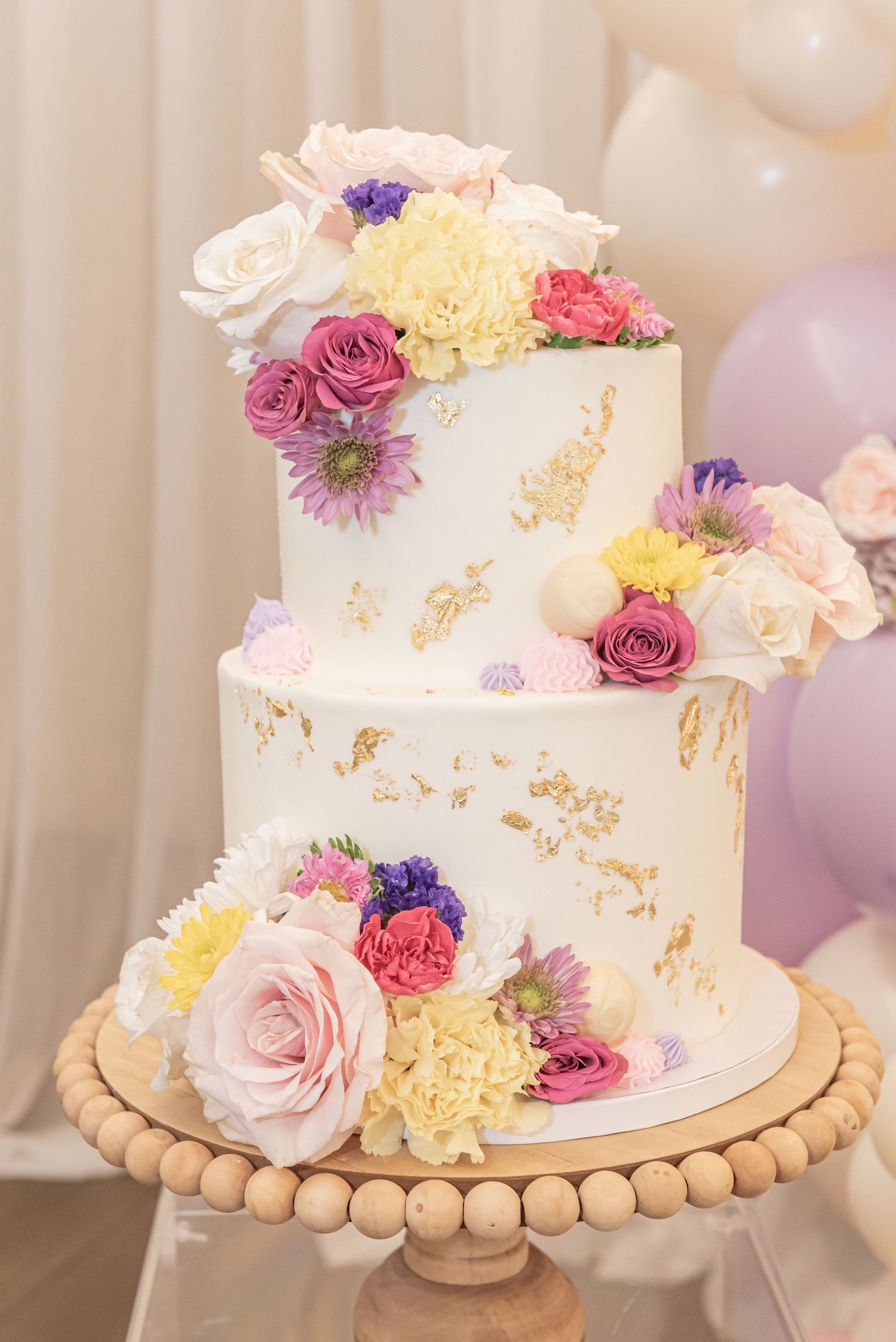 Two-tier white wedding cake decorated with pink, purple and yellow fresh flowers on a wooden beaded cake stand.