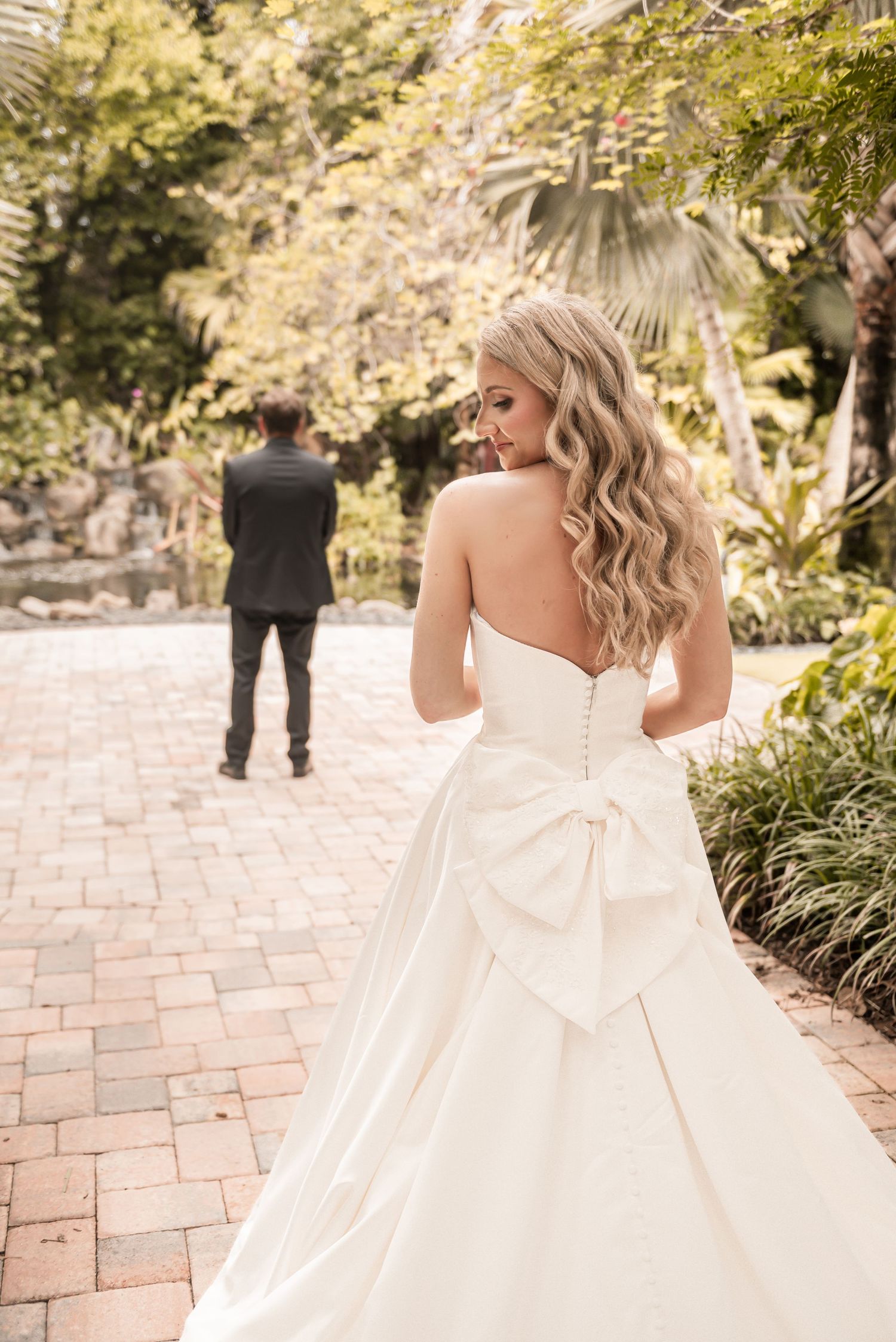 Bride in strapless ball gown with large bow back and wavy blonde hair faces groom in lush garden setting.