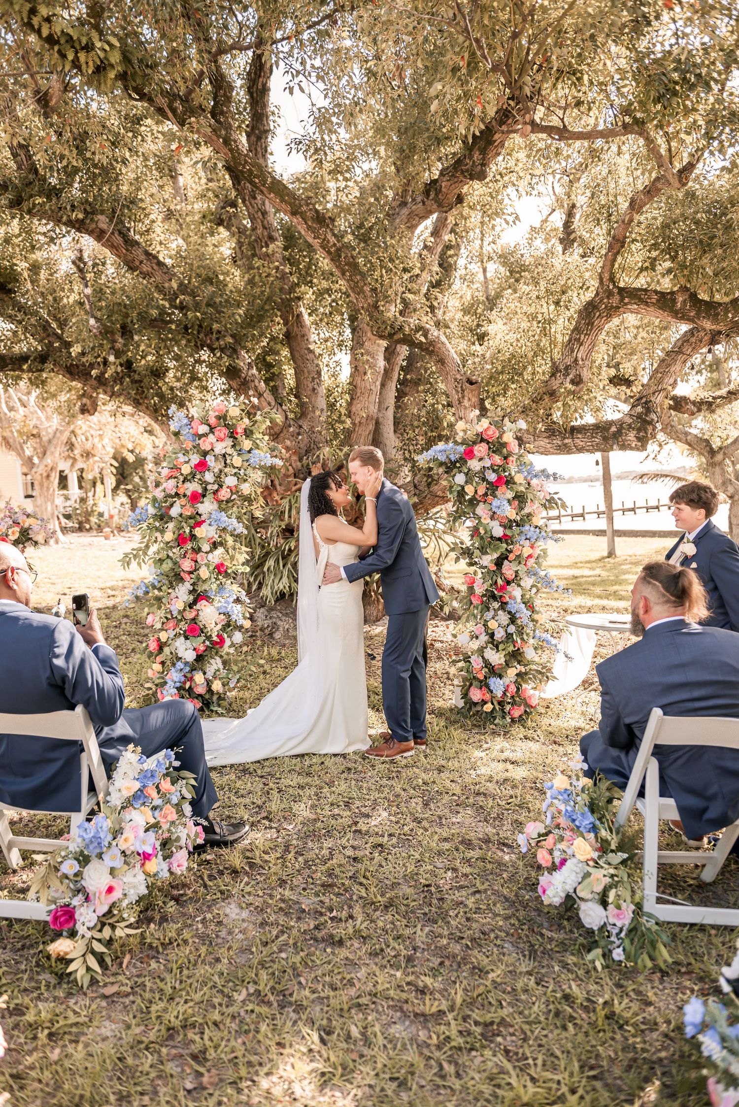 Bride and groom share first kiss under floral arch with pink and blue blooms, surrounded by guests at outdoor wedding ceremony.