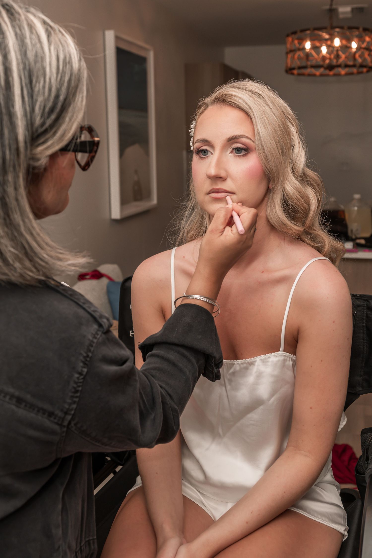 A makeup artist applies finishing touches to a blonde bride in a white slip dress during bridal preparation.