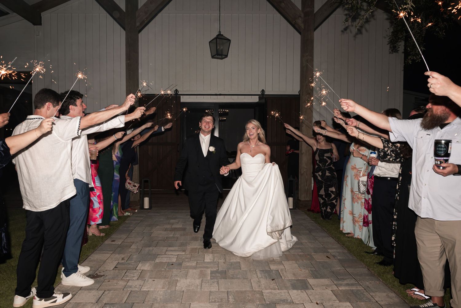 Bride and groom exit wedding venue through sparkler tunnel at night, guests lining the path.