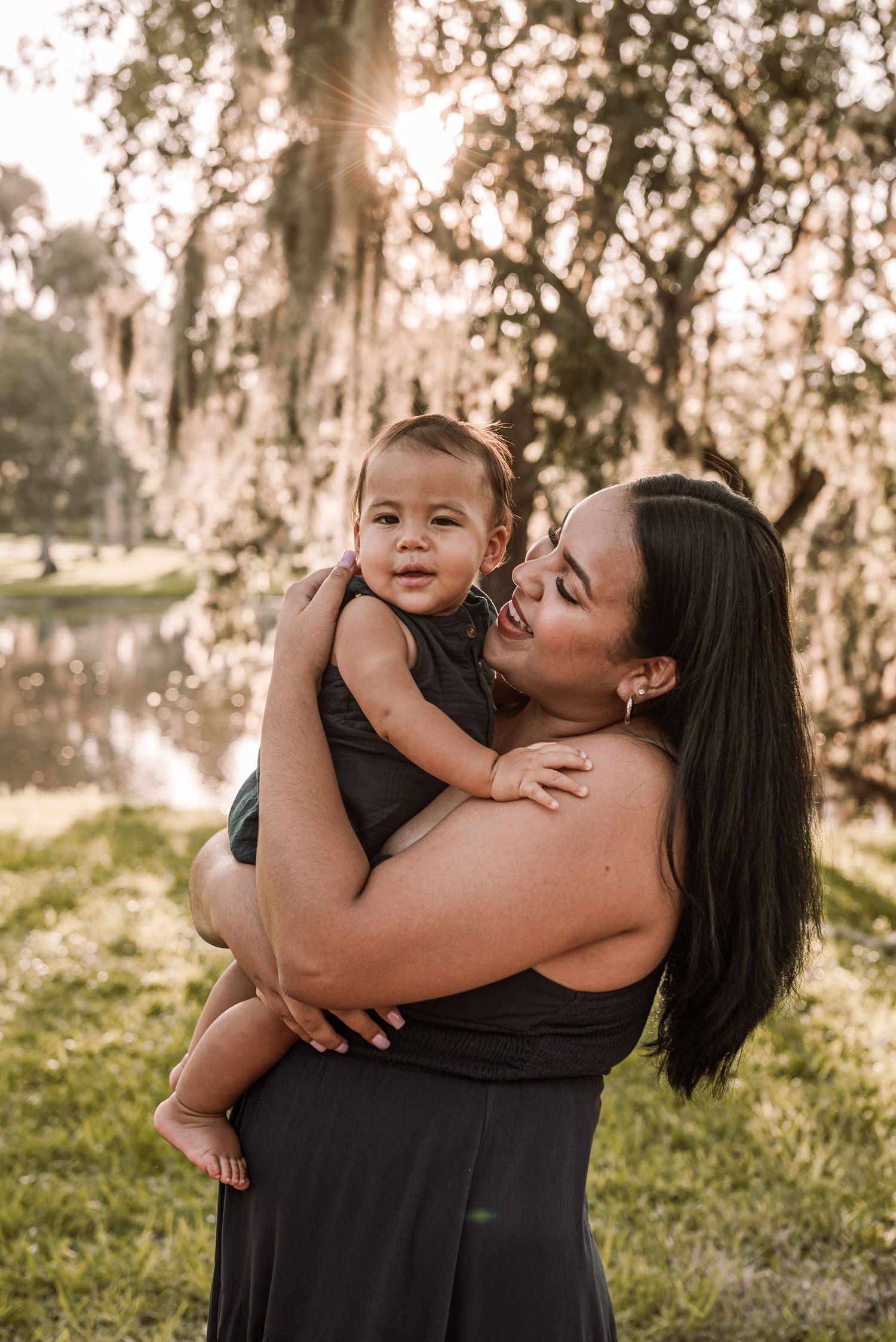 A mother tenderly holds her smiling baby outdoors near a pond with sunlit trees in a warm golden hour setting.