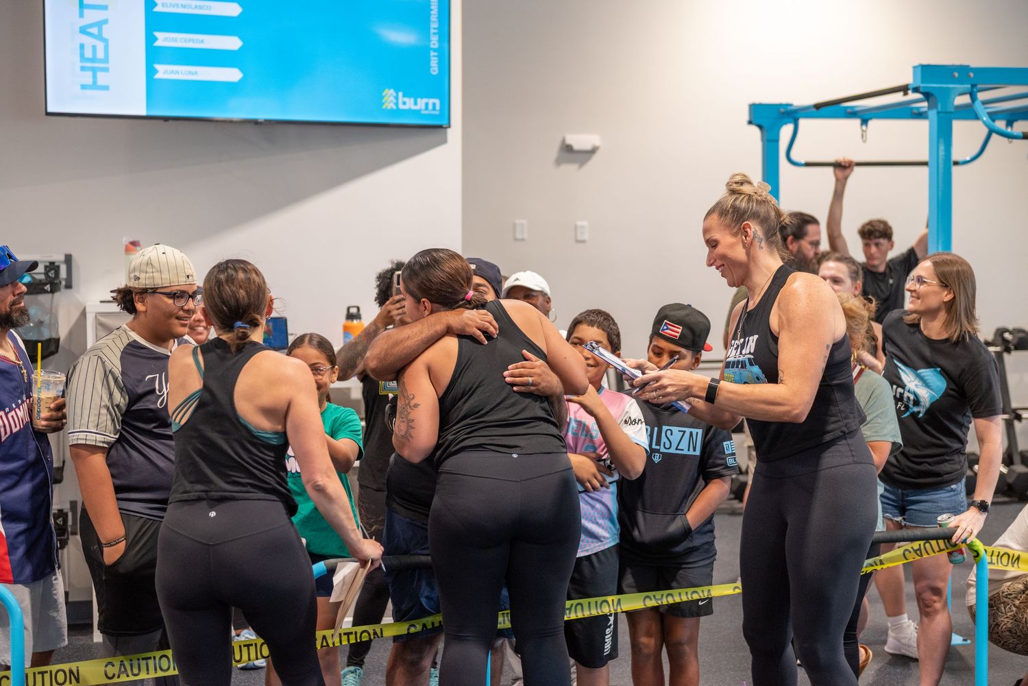 Athletes celebrate together in a gym during a CrossFit competition, hugging and cheering near caution tape barriers.
