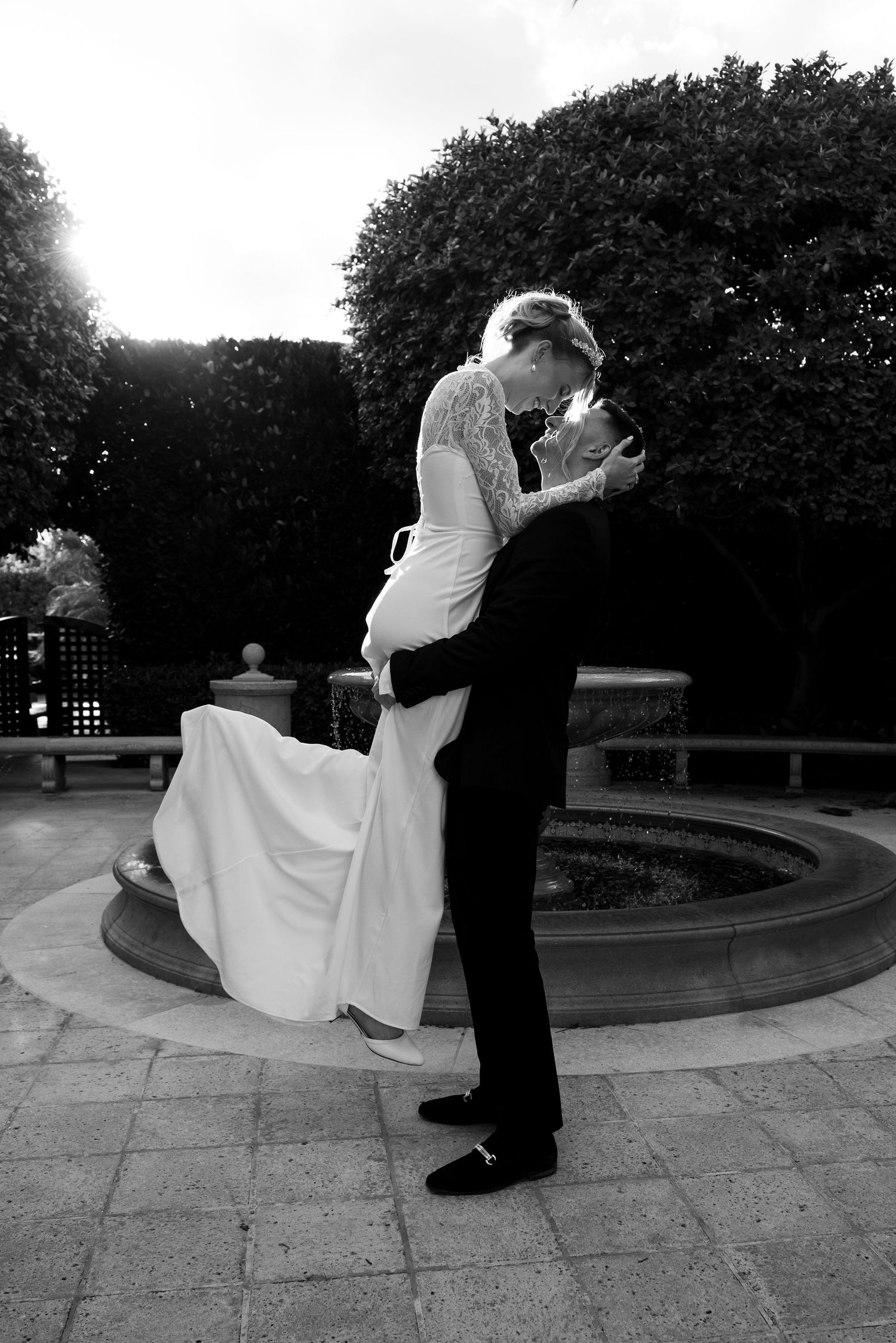 Black and white wedding photo of groom lifting bride near garden fountain, sharing romantic kiss in elegant outdoor setting.