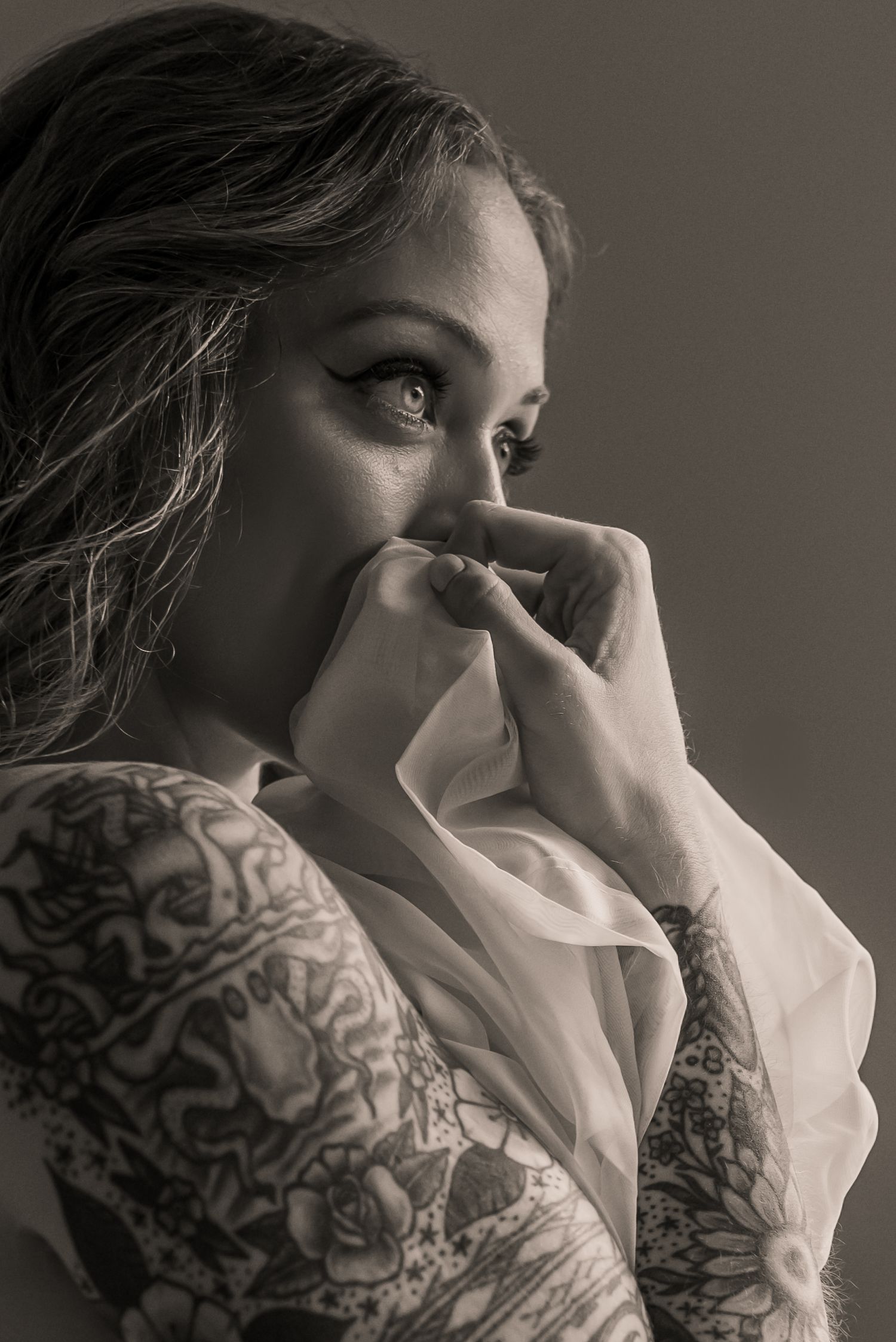 Black and white portrait in soft light with visible arm tattoos and diaphanous fabric covering the mouth.
