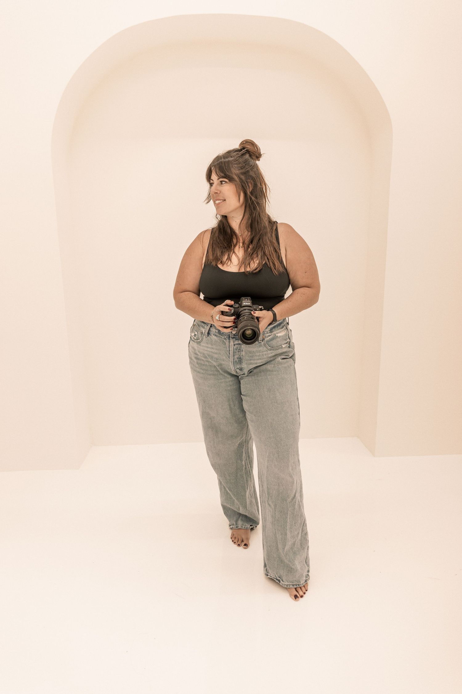 A photographer wearing a brown tank top and light wash jeans poses with a camera in a minimalist white archway.