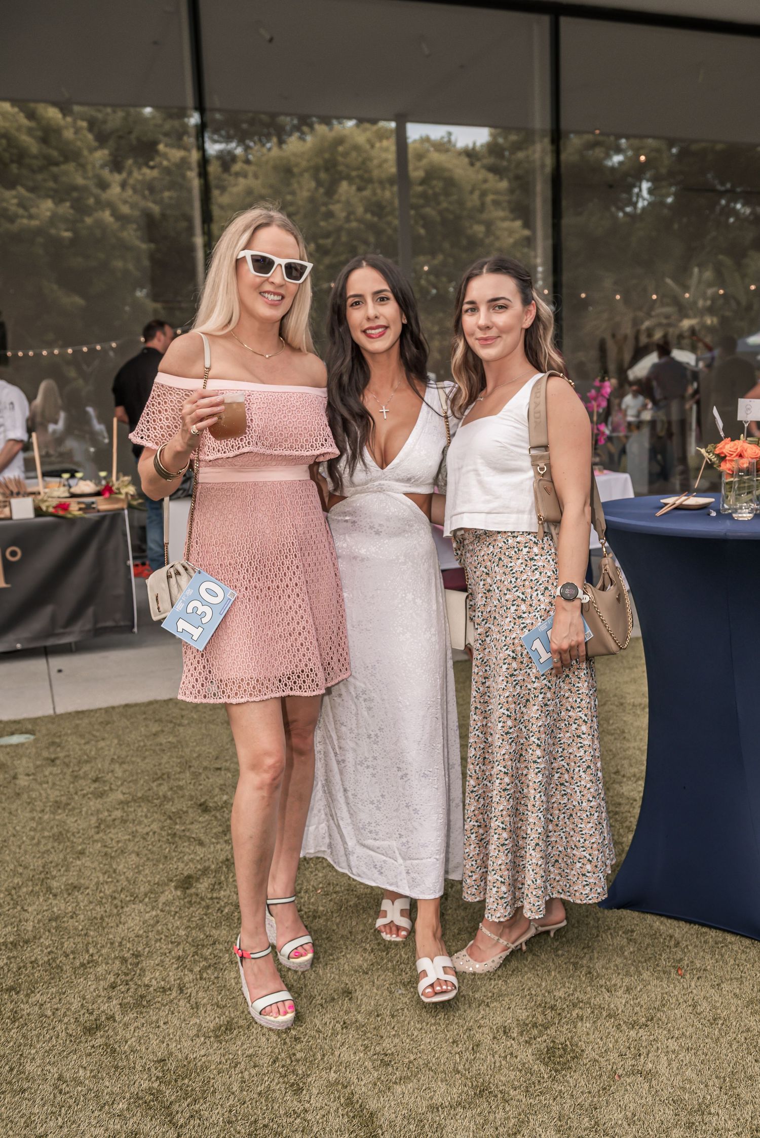 A group of people wearing summer dresses and sandals stand together on grass at an outdoor event.