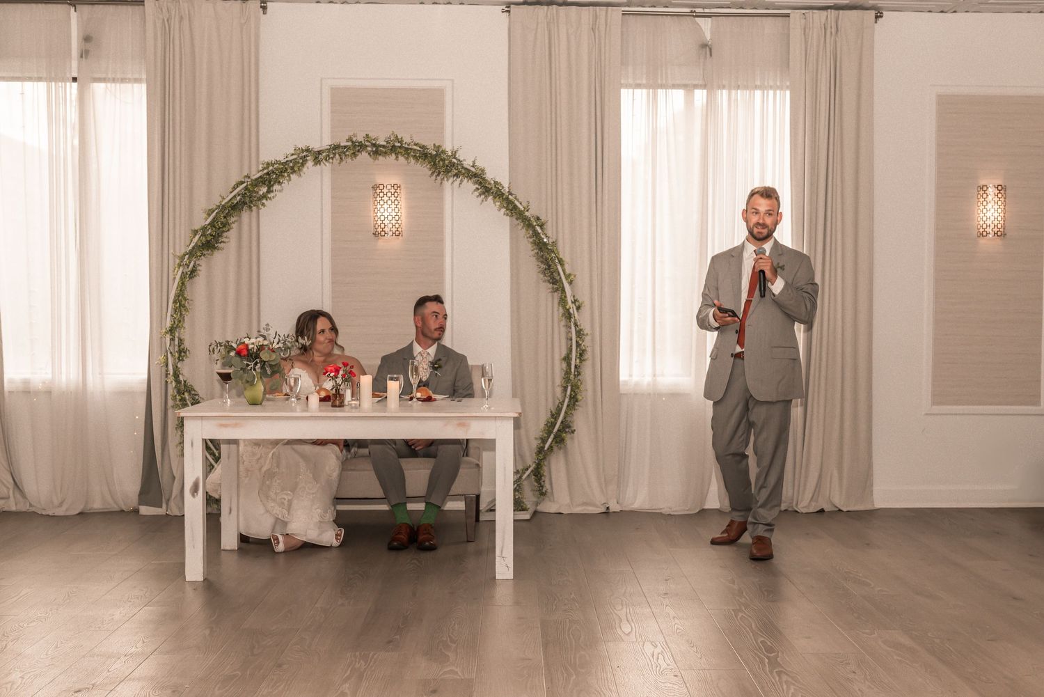 A wedding reception setup featuring an elegant circular greenery arch and head table in a bright venue.