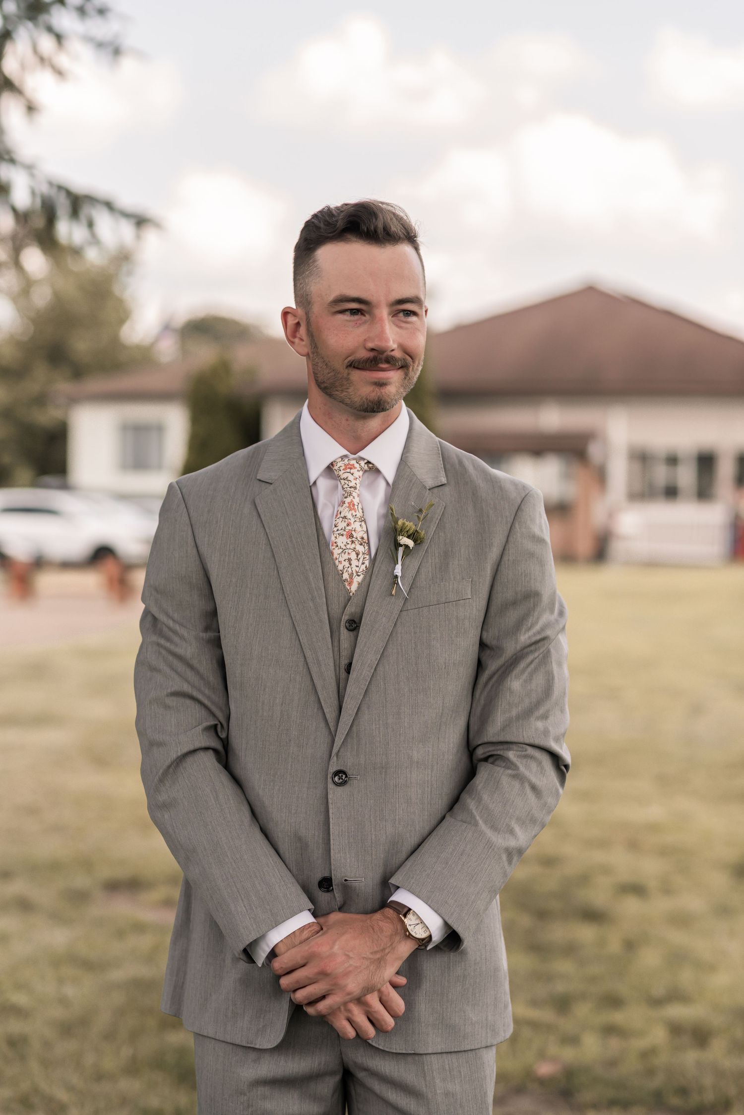 Wedding guest in a light gray three-piece suit with floral tie stands on grass lawn by house on cloudy day.