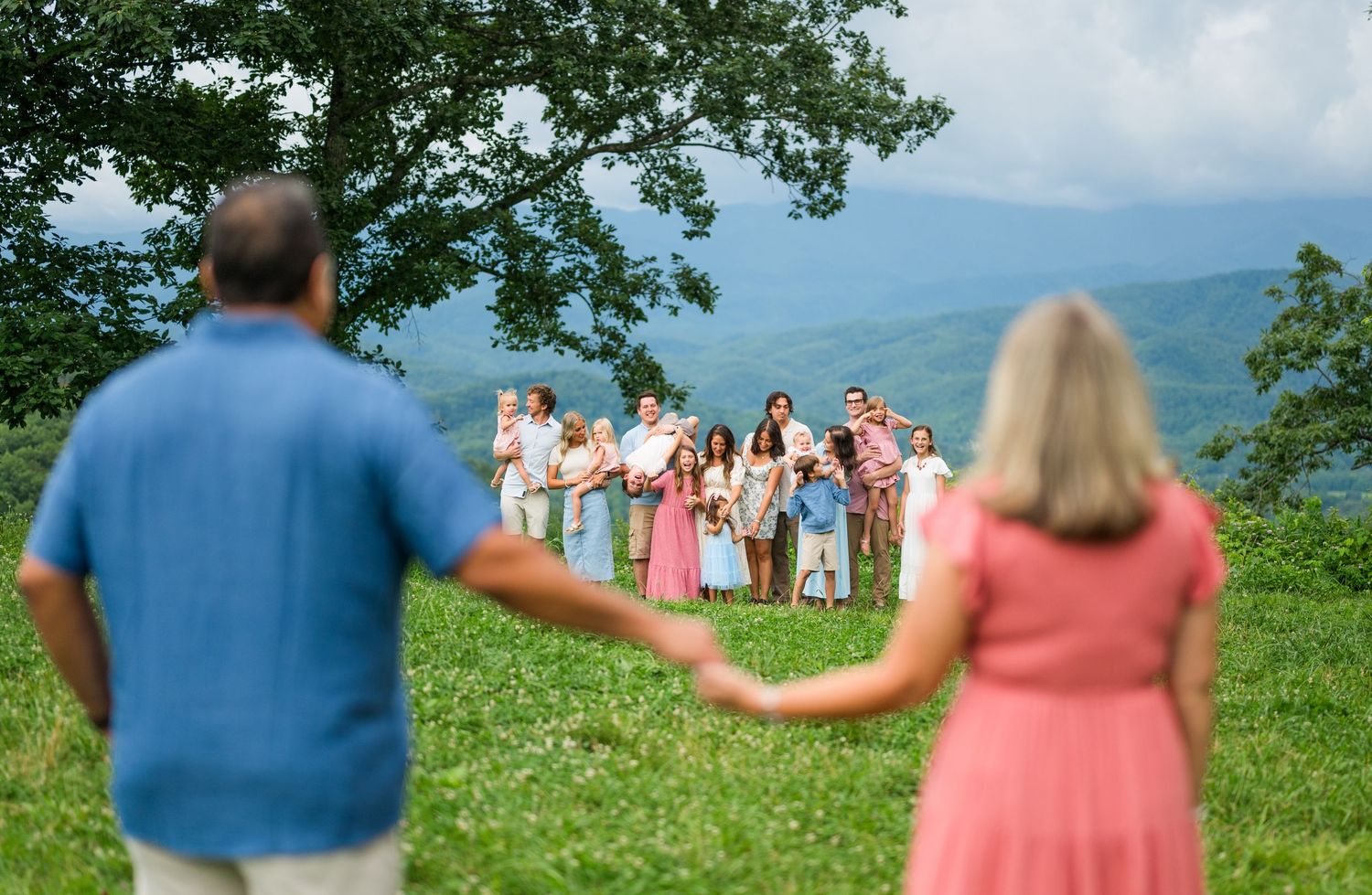 People holding hands and forming a conga line while dancing outdoors on a grassy hillside with trees and mountains in background.