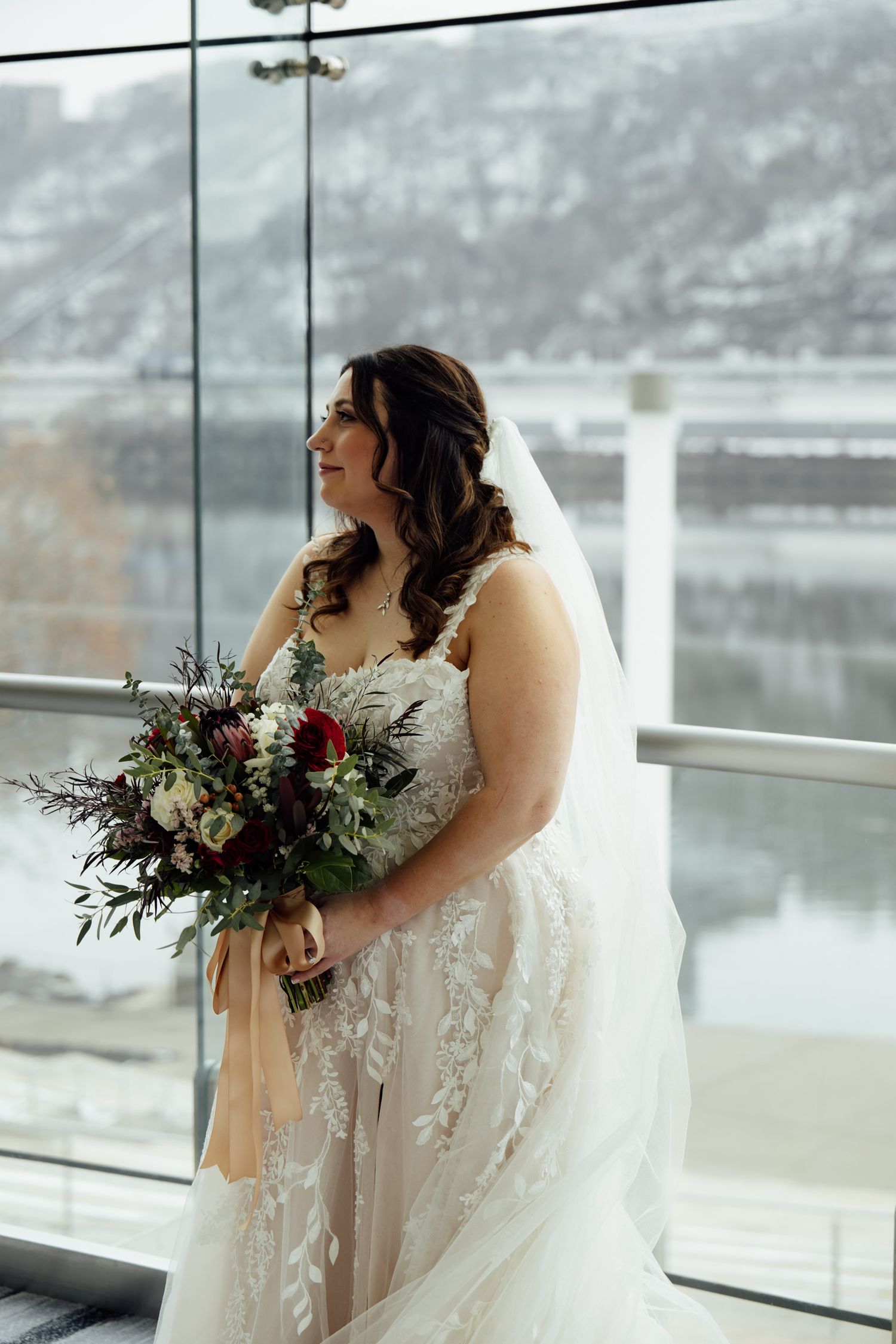 Bride in lace wedding gown holding burgundy and white bouquet stands by glass windows with snowy mountain backdrop.
